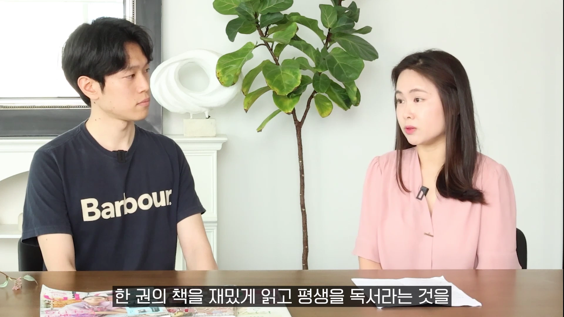 실내에서 남녀 두 사람이 테이블을 사이에 두고 마주 앉아 대화를 나누고 있으며, 뒤쪽에는 화분과 인테리어 소품이 놓여 있습니다.