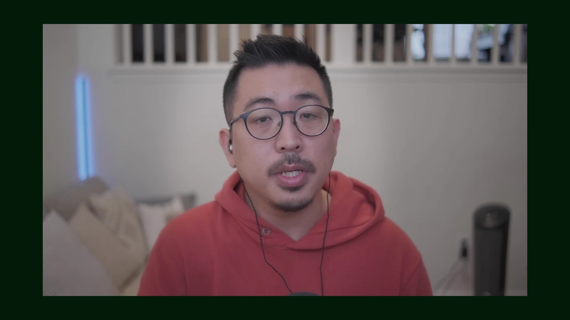 붉은 후드티를 입고 안경을 쓴 남성이 실내에서 카메라를 정면으로 응시하며 이야기하고 있는 모습