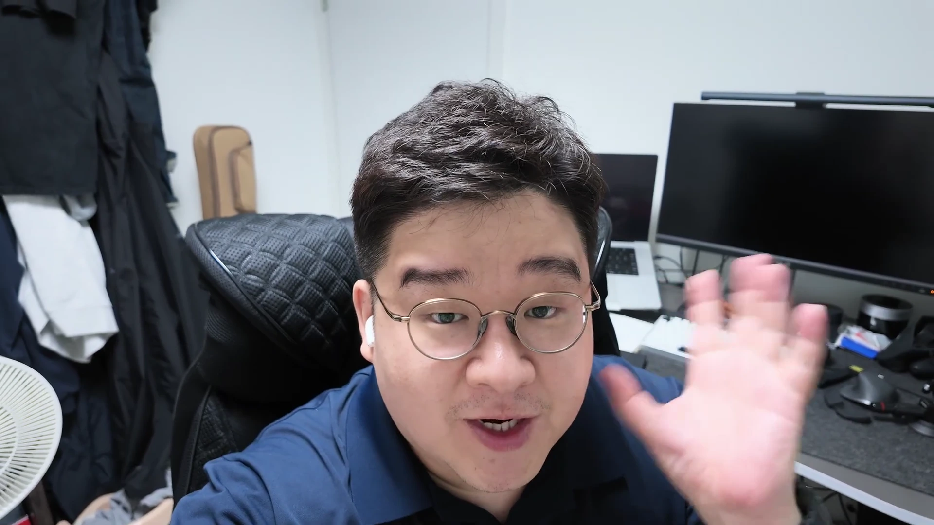 안경을 쓴 남성이 사무실 책상에 앉아 무선 이어폰을 착용한 채 카메라를 향해 손을 흔들며 이야기하고 있다.