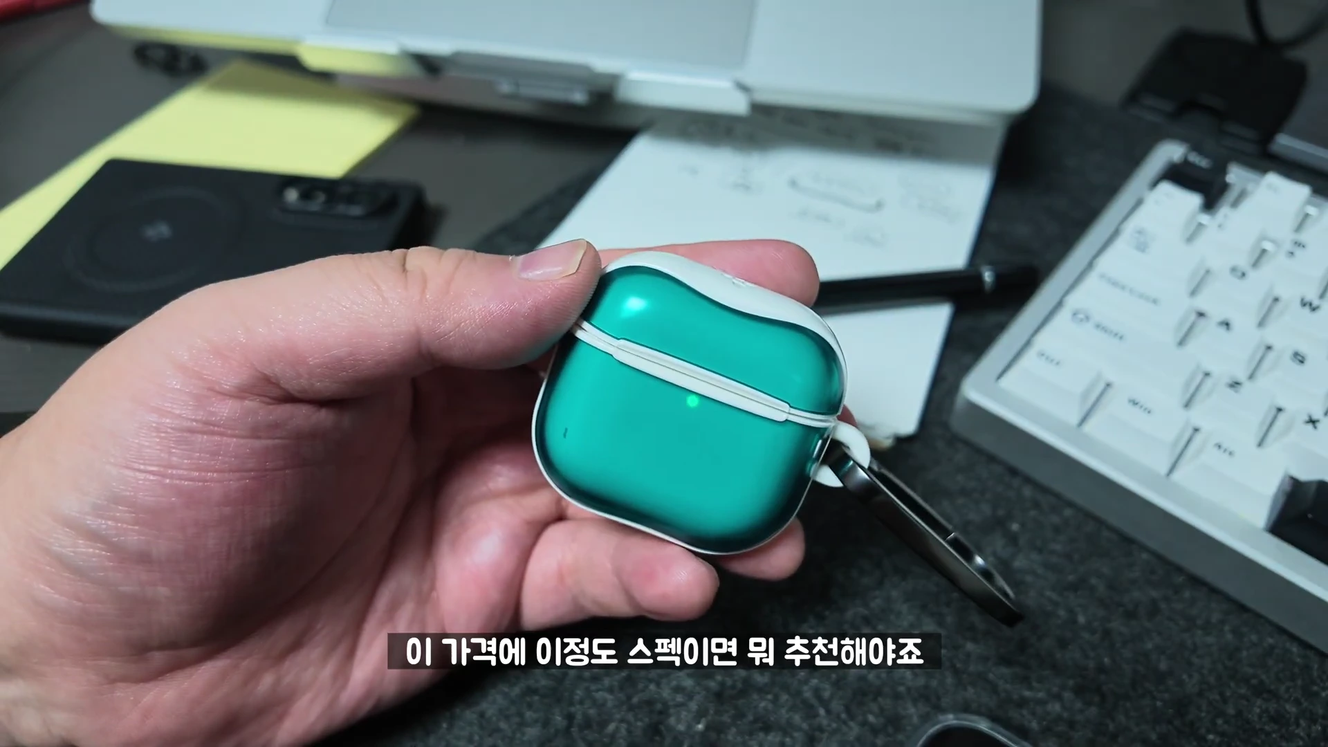손에 들린 청록색 투명 케이스가 씌워진 에어팟 충전 케이스. 책상 위에는 노트북과 키보드가 놓여 있다.