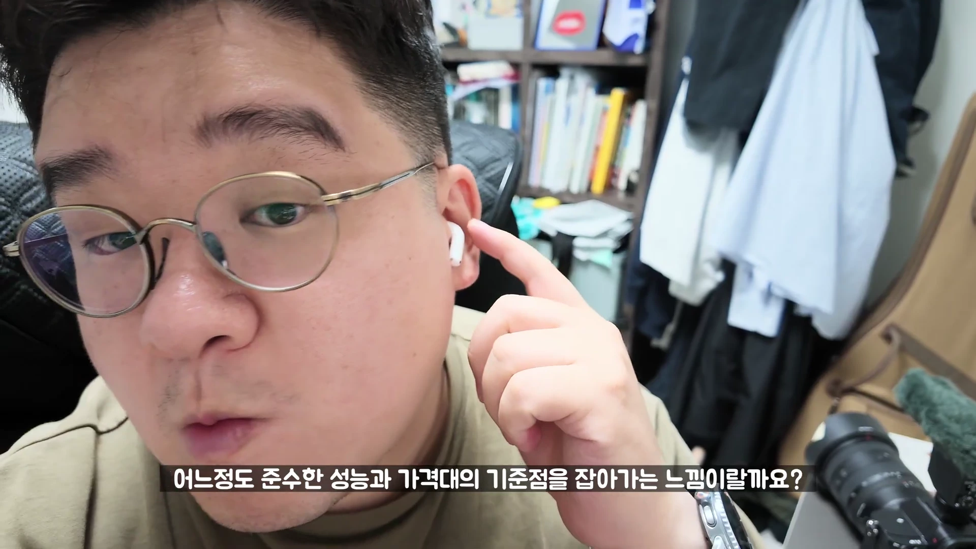 안경을 쓴 남성이 자신의 왼쪽 귀에 오픈형 이어폰을 착용하고 손가락으로 가리키며 카메라를 응시하는 모습