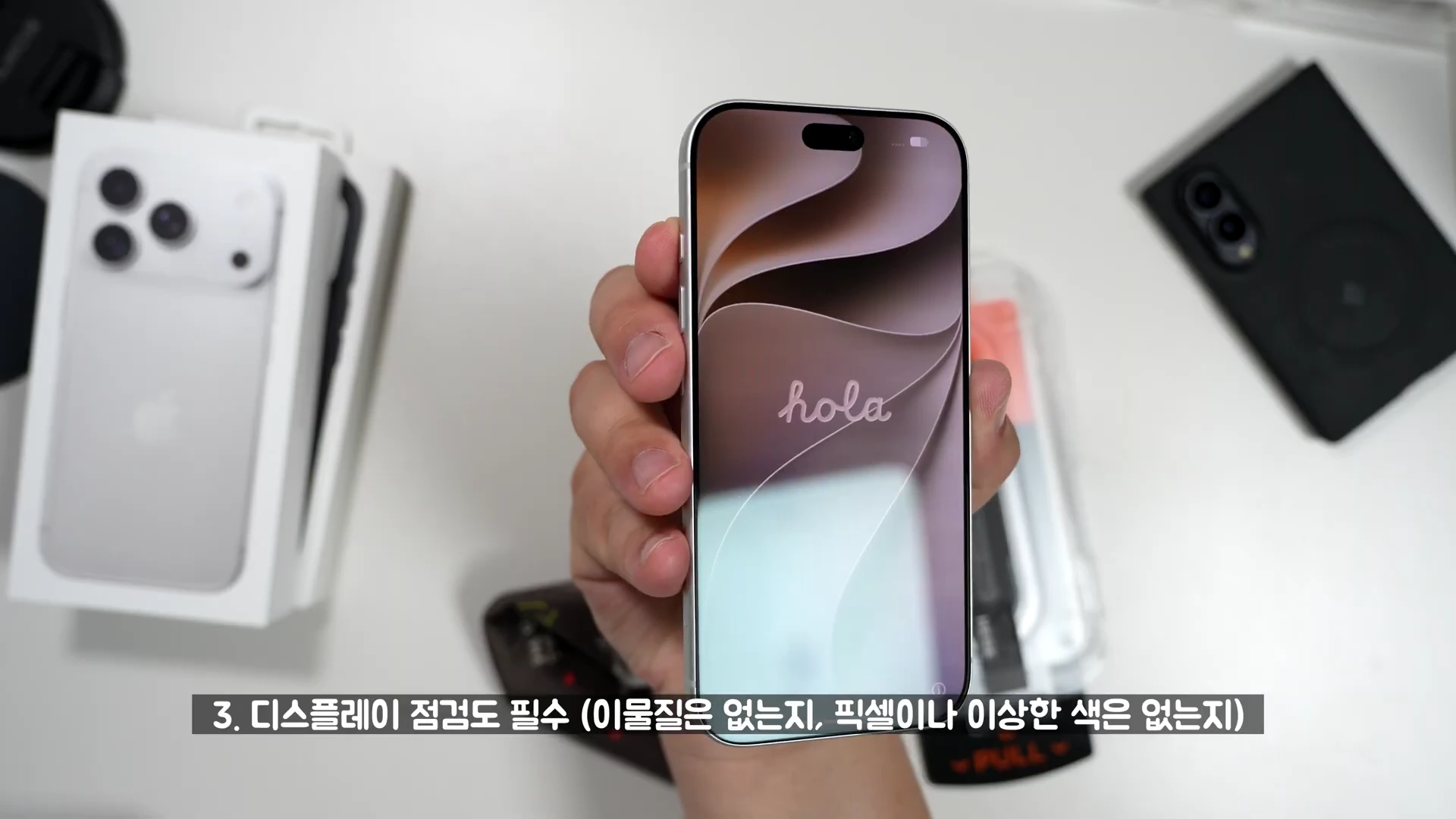 한 손에 들린 아이폰 18 프로 기기와 화면에 'hola'라는 문구가 표시되어 있으며, 하단에 점검 사항에 관한 한국어 자막이 있는 화면.