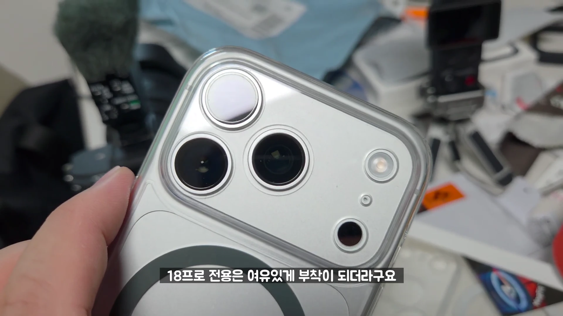 한 손으로 들고 있는 실버 색상의 스마트폰 뒷면 카메라 섬 부분과 그 위에 투명 케이스가 장착된 모습.