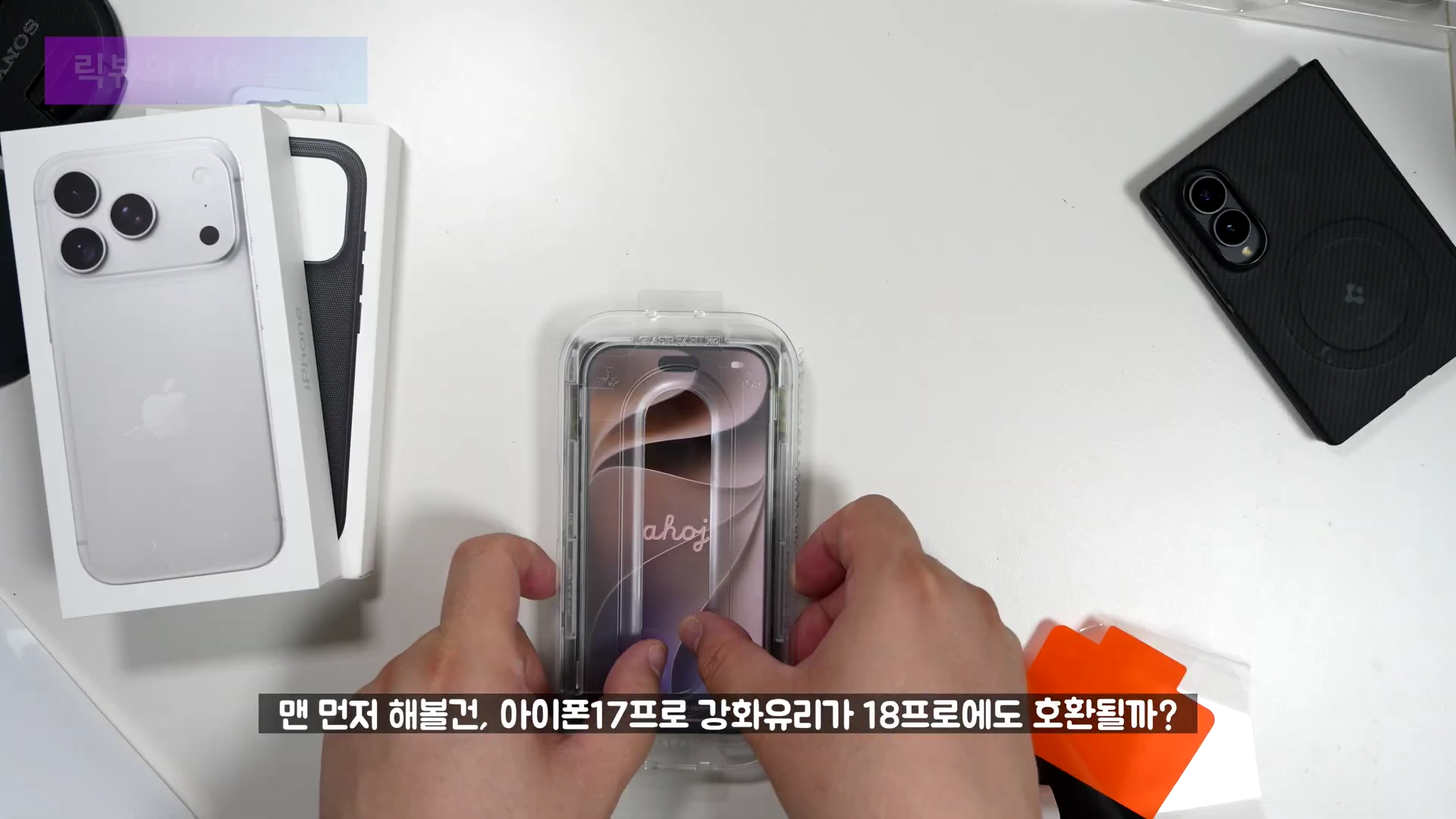 흰색 배경의 책상 위에서 양손으로 아이폰 17용 강화유리 부착 도구를 조립하고 있는 모습이 담긴 항공샷.