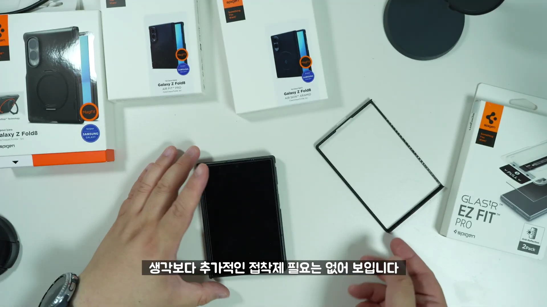 밝은 테이블 위에 갤럭시 폴드8용 슈피겐 케이스 제품 상자들과 스마트폰, 그리고 케이스 부품이 놓여 있고 사람의 손이 이를 조립하고 있는 모습