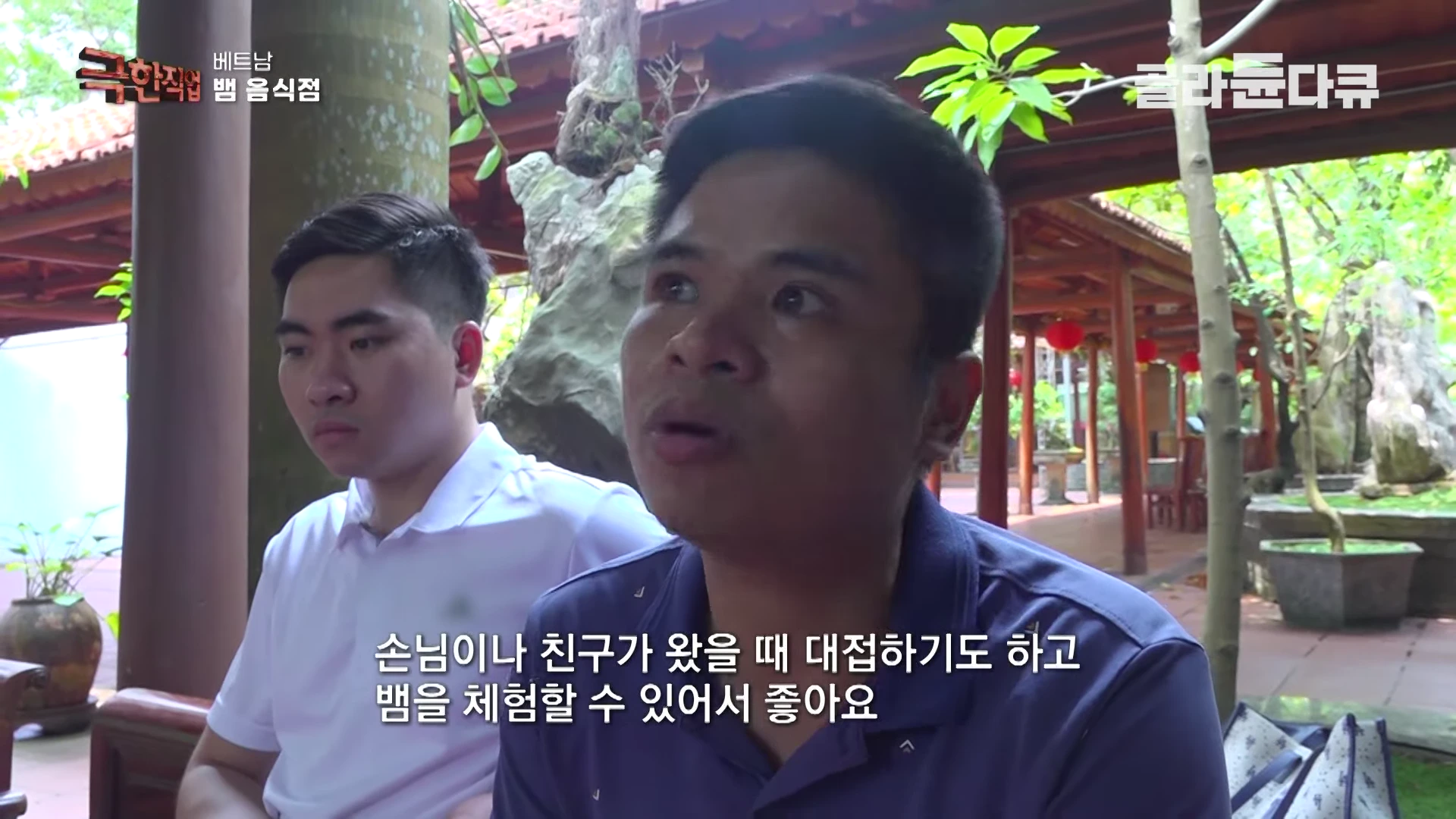 실내 식당에 앉아 있는 두 명의 베트남 남성들이 카메라를 향해 대화를 나누고 있다.