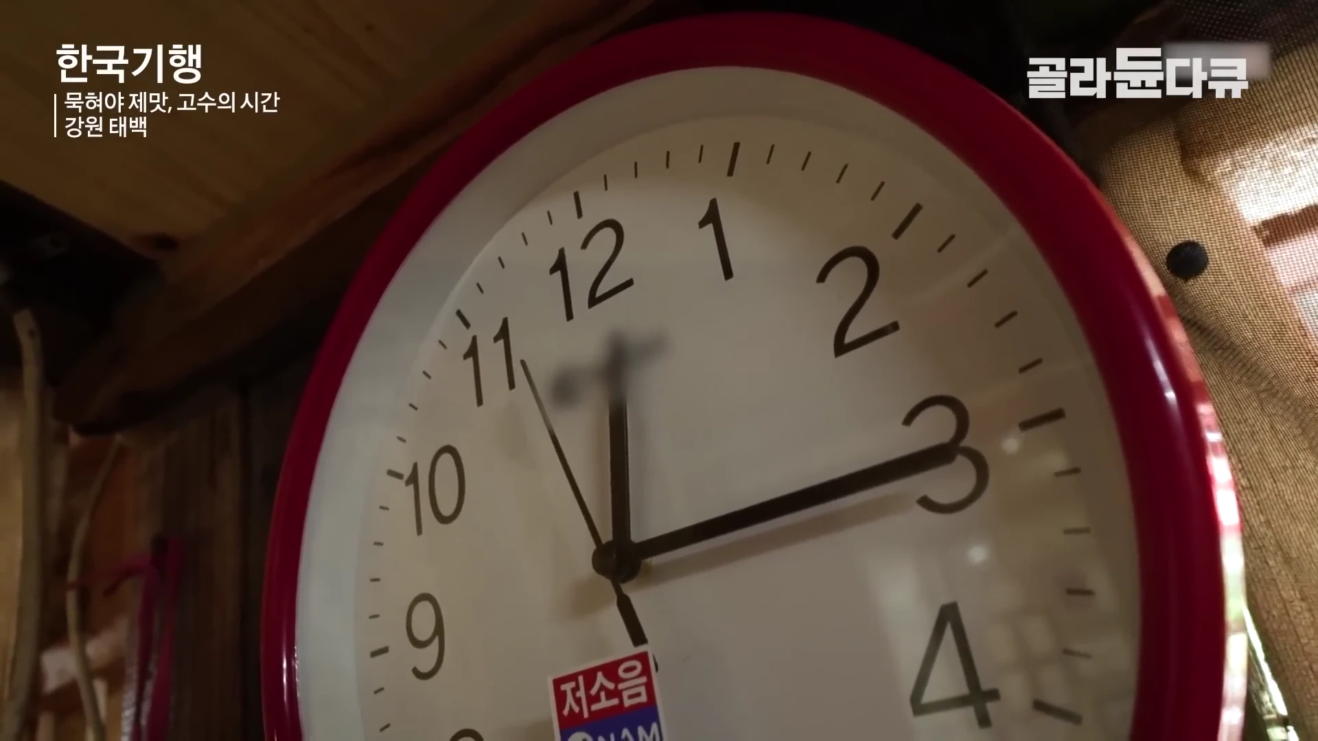 붉은색 테두리를 가진 벽걸이 시계가 12시 15분쯤을 가리키고 있는 클로즈업 화면입니다.