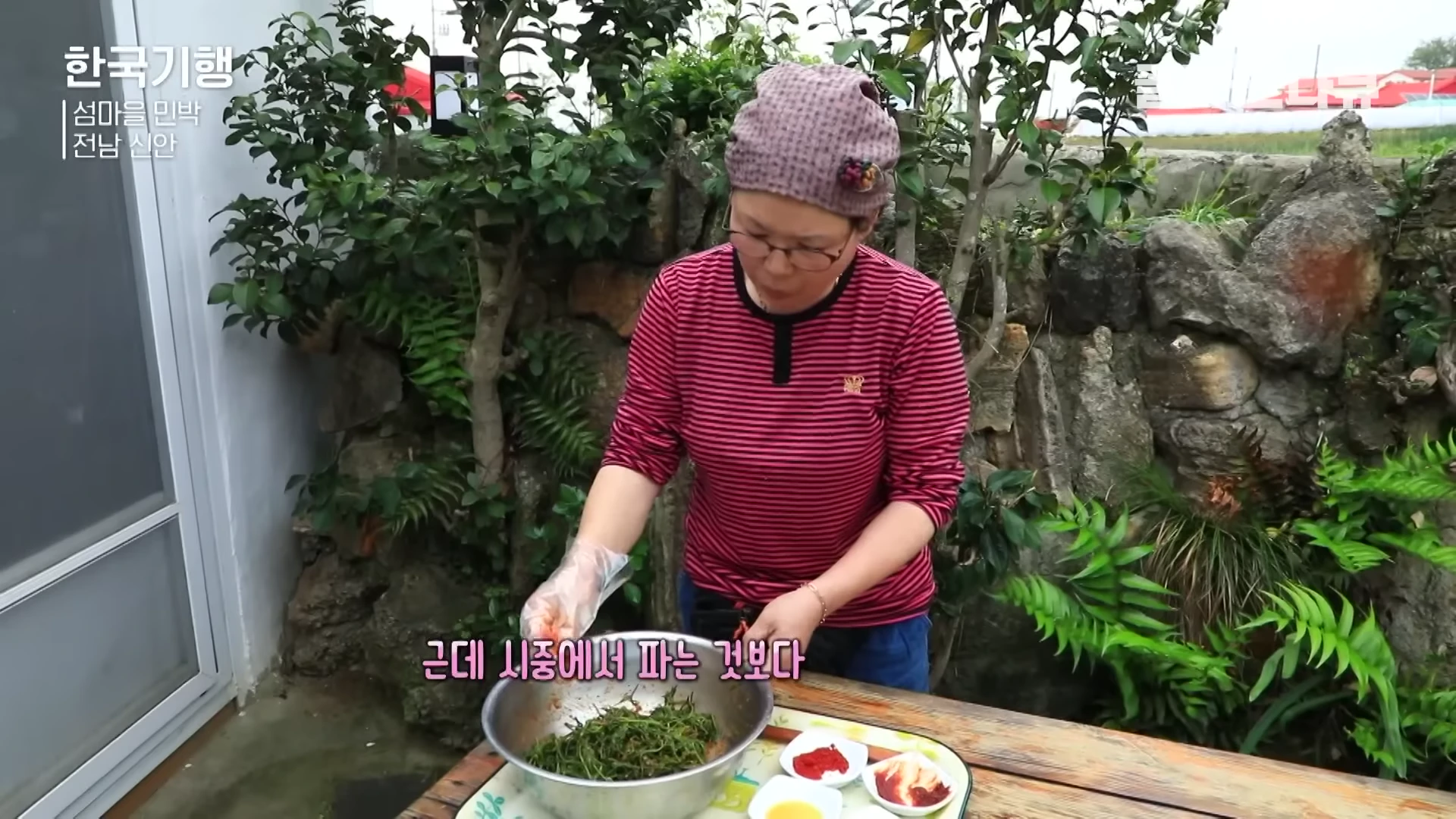 한 여성이 마당에서 비닐장갑을 끼고 양푼에 담긴 나물을 손으로 정성스럽게 무치고 있는 모습