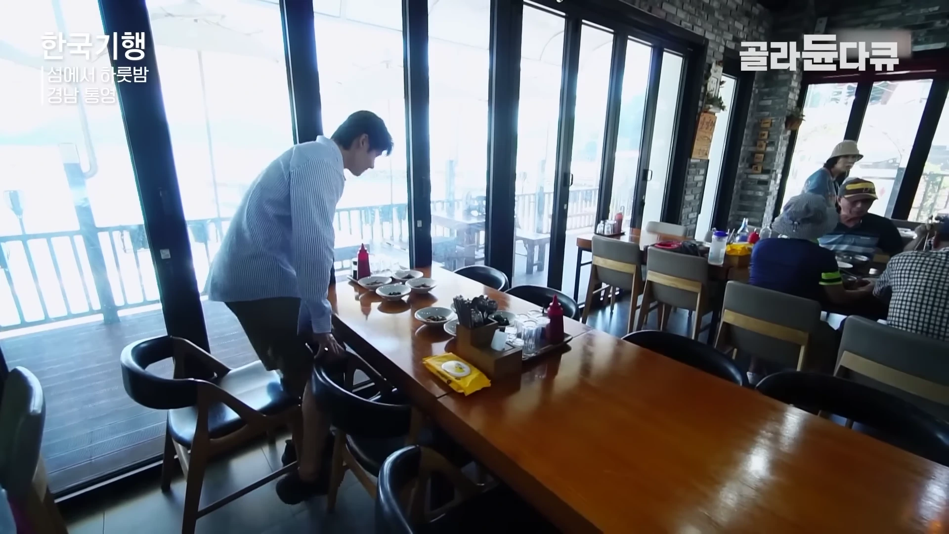 섬마을 민박 식당 안에서 한 남성이 긴 식탁 옆에 서 있고 뒤쪽 좌석에는 여러 명의 손님이 앉아 식사를 하고 있는 모습