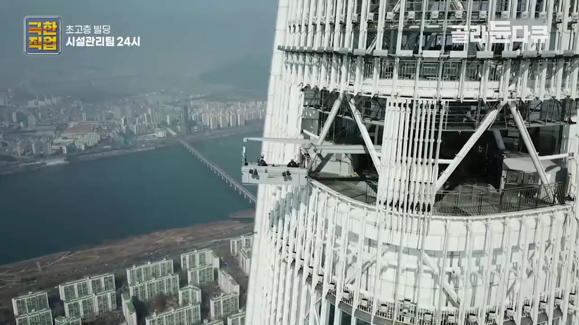 초고층 빌딩 외벽에 매달린 곤돌라에 작업자들이 탑승하여 유지보수 작업을 수행하고 있으며 그 아래로 한강과 도시 풍경이 내려다보이는 공중 촬영 장면