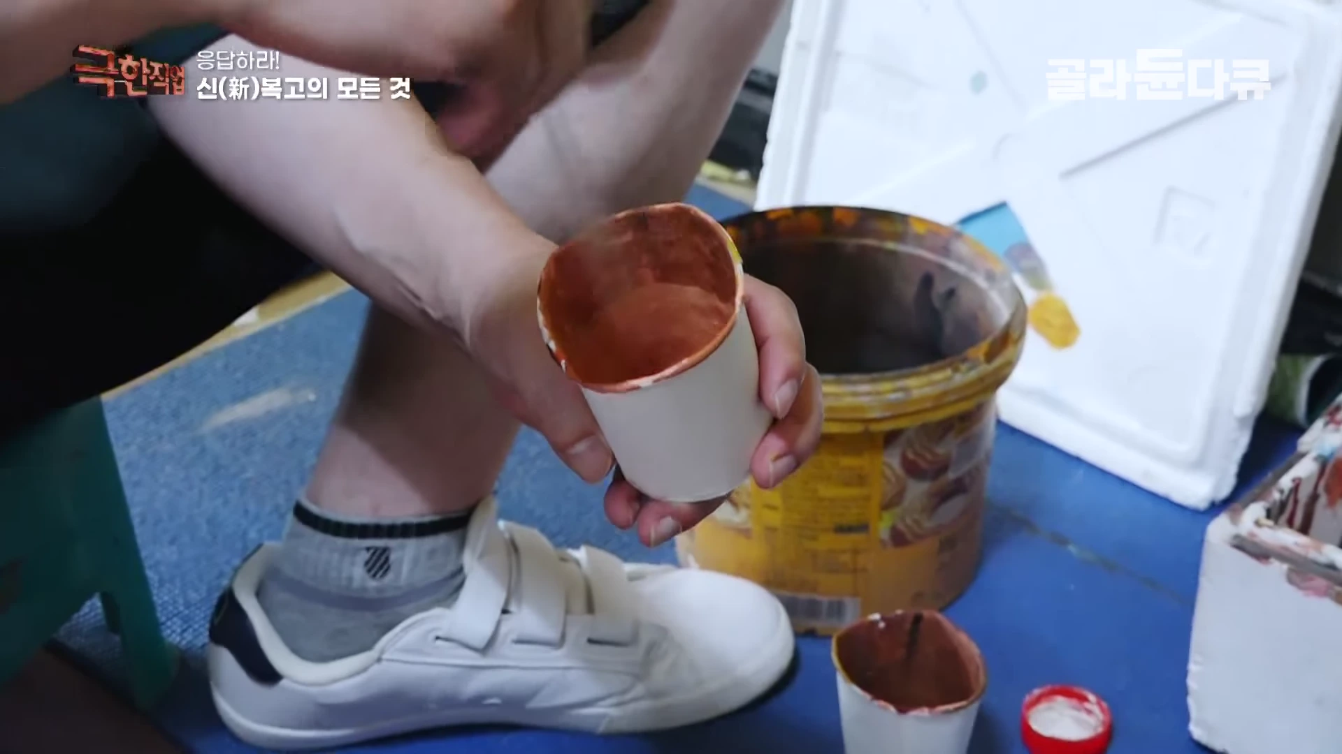 작업자가 종이컵에 든 페인트를 손에 들고 옆에 놓인 큰 페인트 통과 함께 작업실 바닥에 앉아 있는 모습