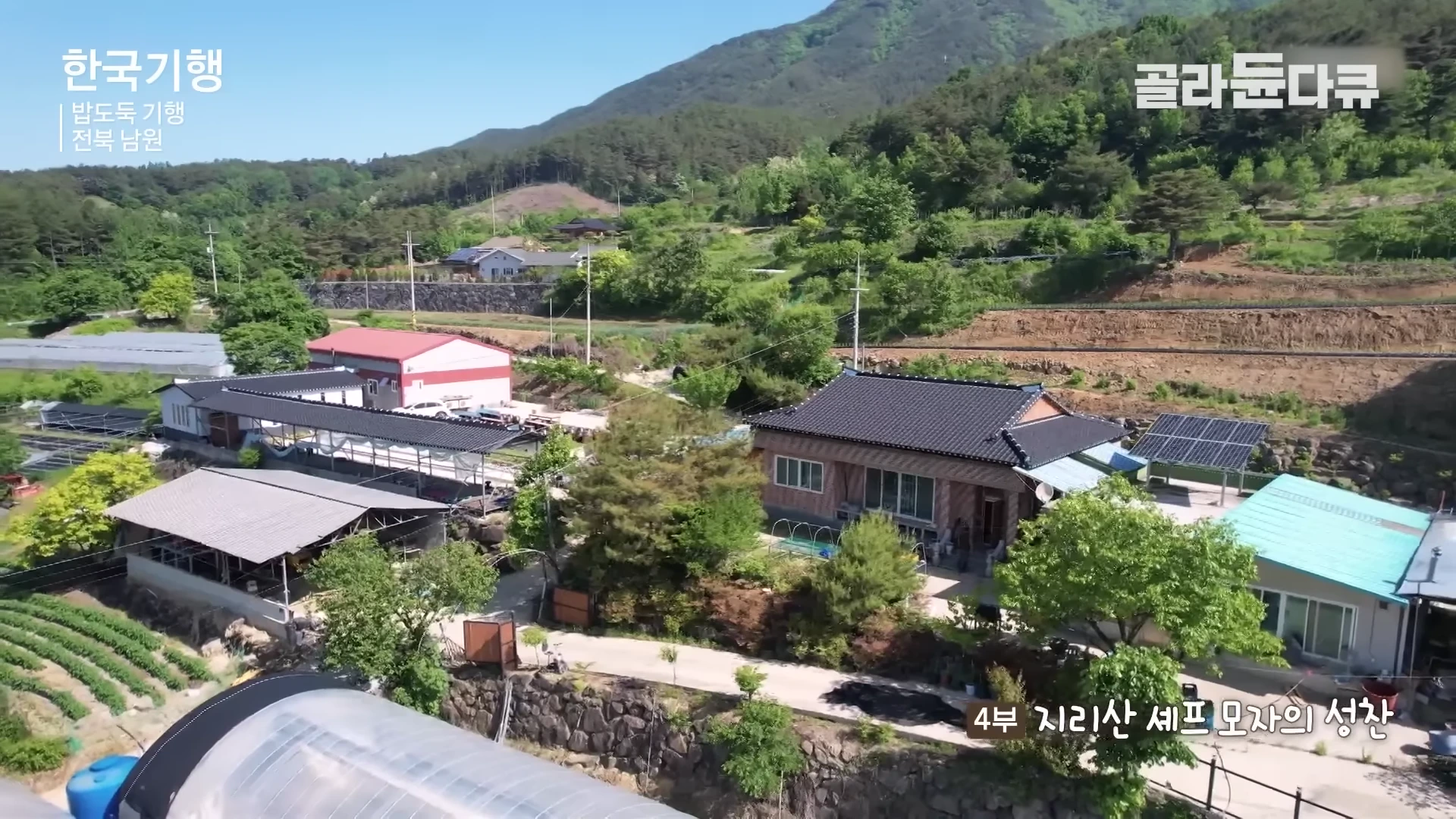 푸른 산으로 둘러싸인 지리산 자락의 시골 마을 전경이 드론으로 내려다보이며, 기와지붕의 단독 주택과 창고들이 보입니다.
