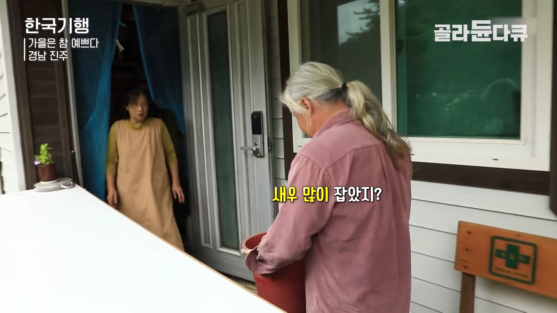 저수지 인근 집 현관 앞에서 긴 머리를 뒤로 묶은 남성이 빨간색 양동이를 들고 있고, 그 뒤로 앞치마를 두른 여성이 서 있는 모습