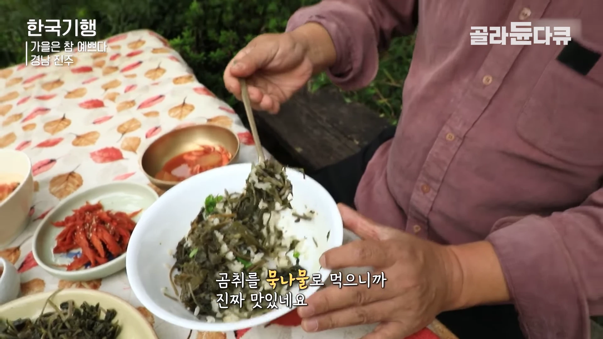 갈색 셔츠를 입은 남성이 숟가락으로 곰치 묵나물을 밥 위에 얹고 있는 모습이 담긴 클로즈업 화면입니다. 식탁 위에는 다양한 나물 반찬이 놓여 있습니다.