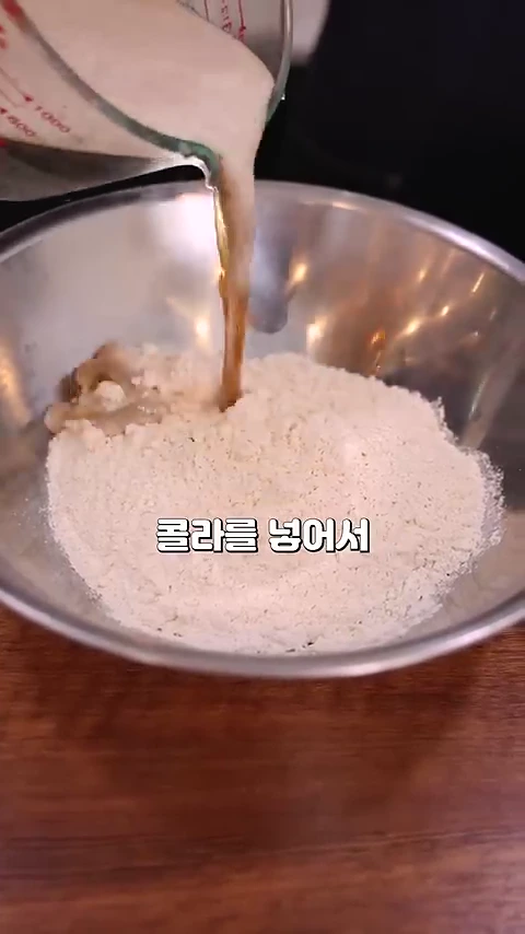 투명한 계량컵에 든 갈색의 콜라를 금속 볼에 담긴 하얀 튀김가루 위로 붓는 모습이며, 화면 중앙 하단에 '콜라를 넣어서'라는 한국어 자막이 표시되어 있습니다.