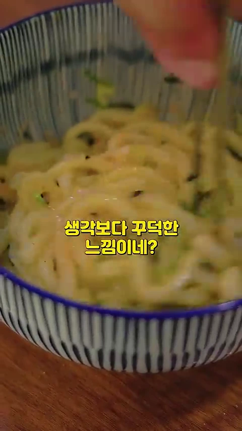 젓가락으로 비벼지고 있는 명란 가마타마 우동이 담긴 줄무늬 그릇의 근접 촬영 화면. 화면 중앙에는 '생각보다 꾸덕한 느낌이네?'라는 자막이 노란색으로 적혀 있다.
