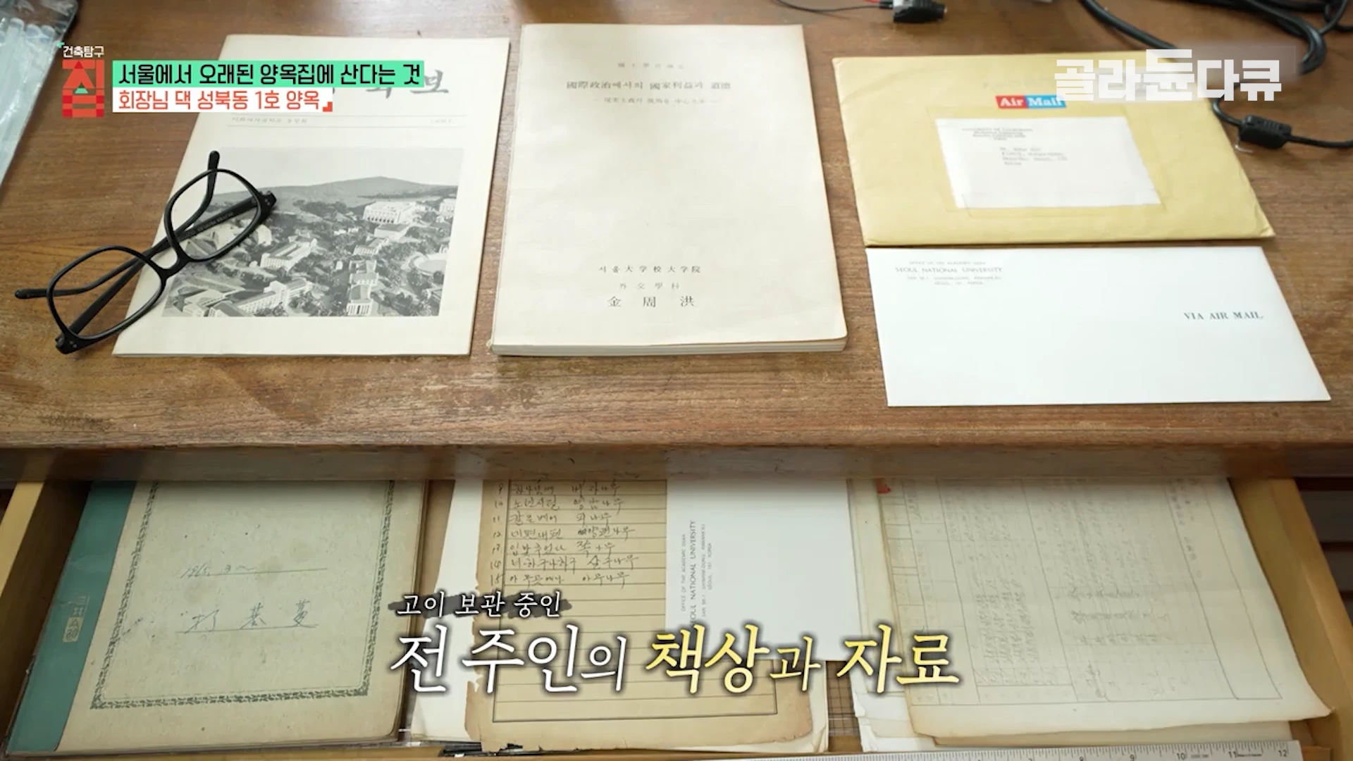 나무 책상 위에 안경과 여러 장의 낡은 서류, 항공 우편 봉투가 놓여 있고, 열린 서랍 속에는 정갈하게 정리된 기록물들이 보인다.