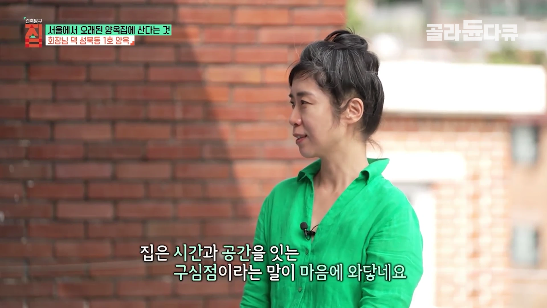 붉은 벽돌 벽을 배경으로 초록색 셔츠를 입은 여성이 측면을 바라보며 인터뷰하는 모습이며 하단에 한글 자막이 있다.
