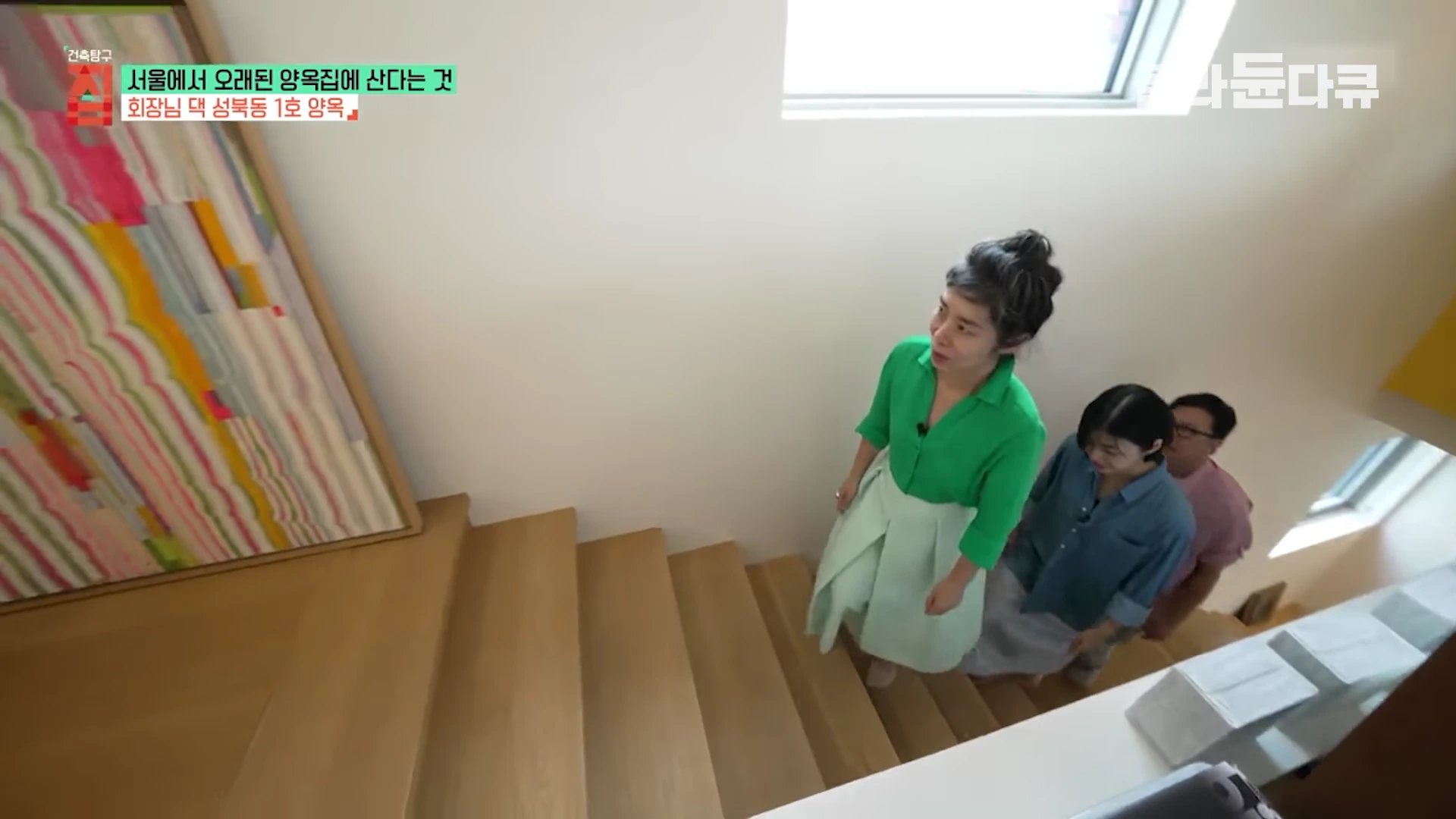 실내 계단을 오르는 세 명의 사람들이 보이고, 벽면에는 추상화 작품이 걸려 있으며 계단 옆으로는 유리 난간이 설치되어 있다.