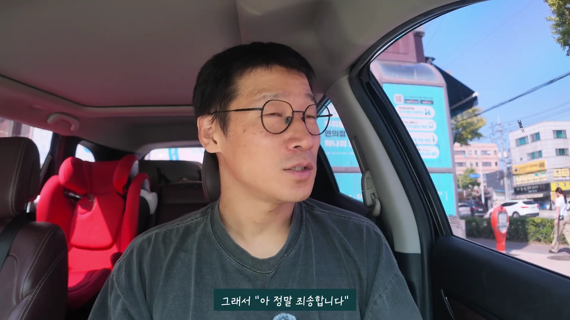 안경을 쓴 남성이 자동차 운전석에 앉아 정면을 보며 이야기를 하고 있으며, 뒷좌석에는 빨간색 어린이용 카시트가 놓여 있다.