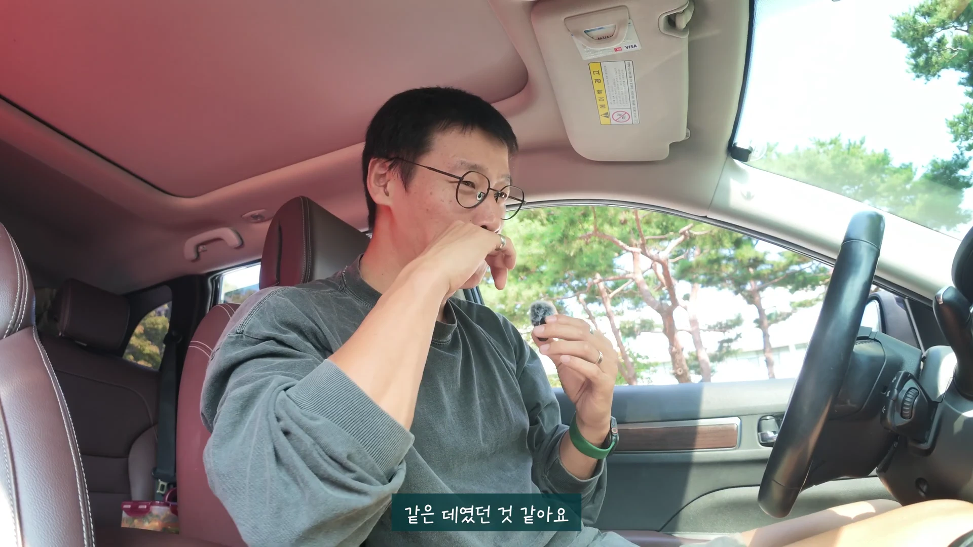 안경을 쓴 남성이 자동차 운전석에 앉아 마이크를 손에 쥐고 이야기하고 있으며 화면 하단에는 자막이 있다.