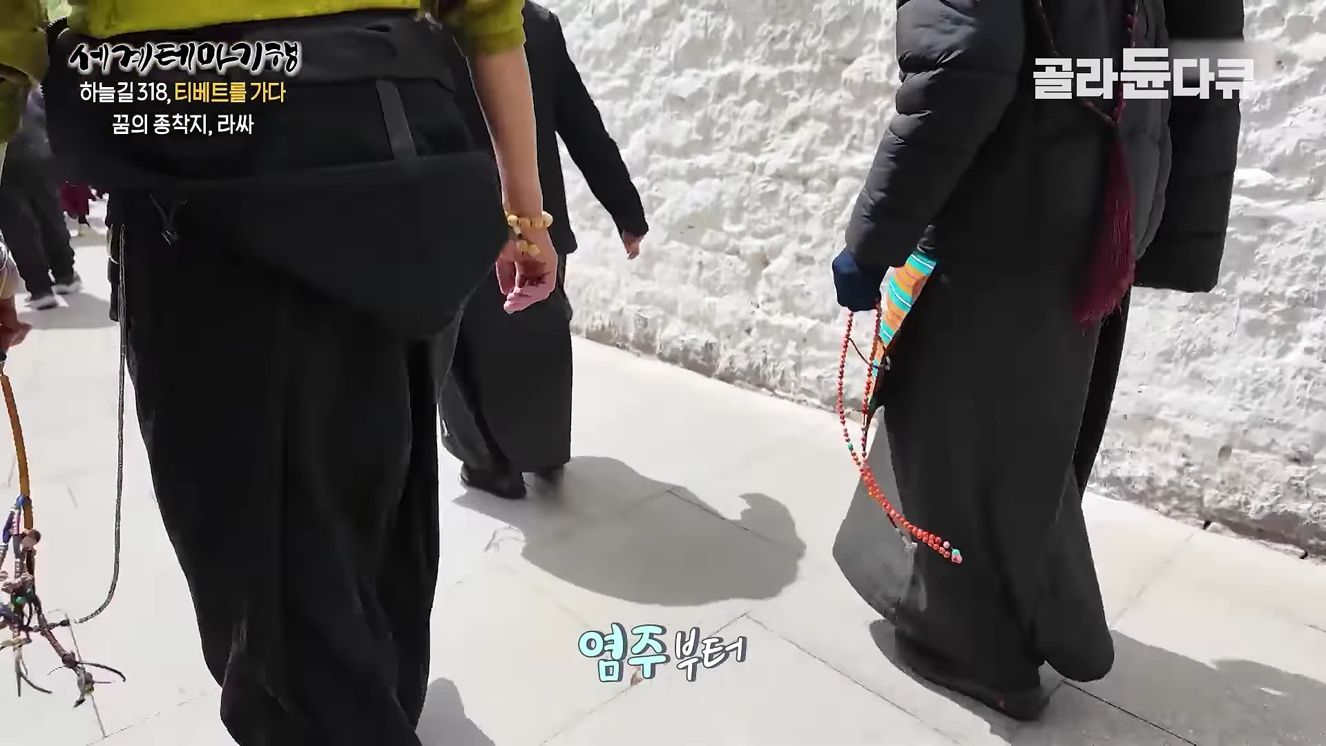 흰색 외벽을 따라 걷는 여러 명의 순례자들의 뒷모습으로, 이들은 손에 염주나 기도 도구를 들고 있다.
