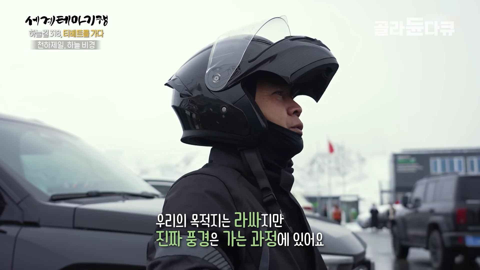 오토바이 헬멧을 쓰고 있는 남성이 흐린 배경의 설산 근처에서 어딘가를 바라보고 있다.