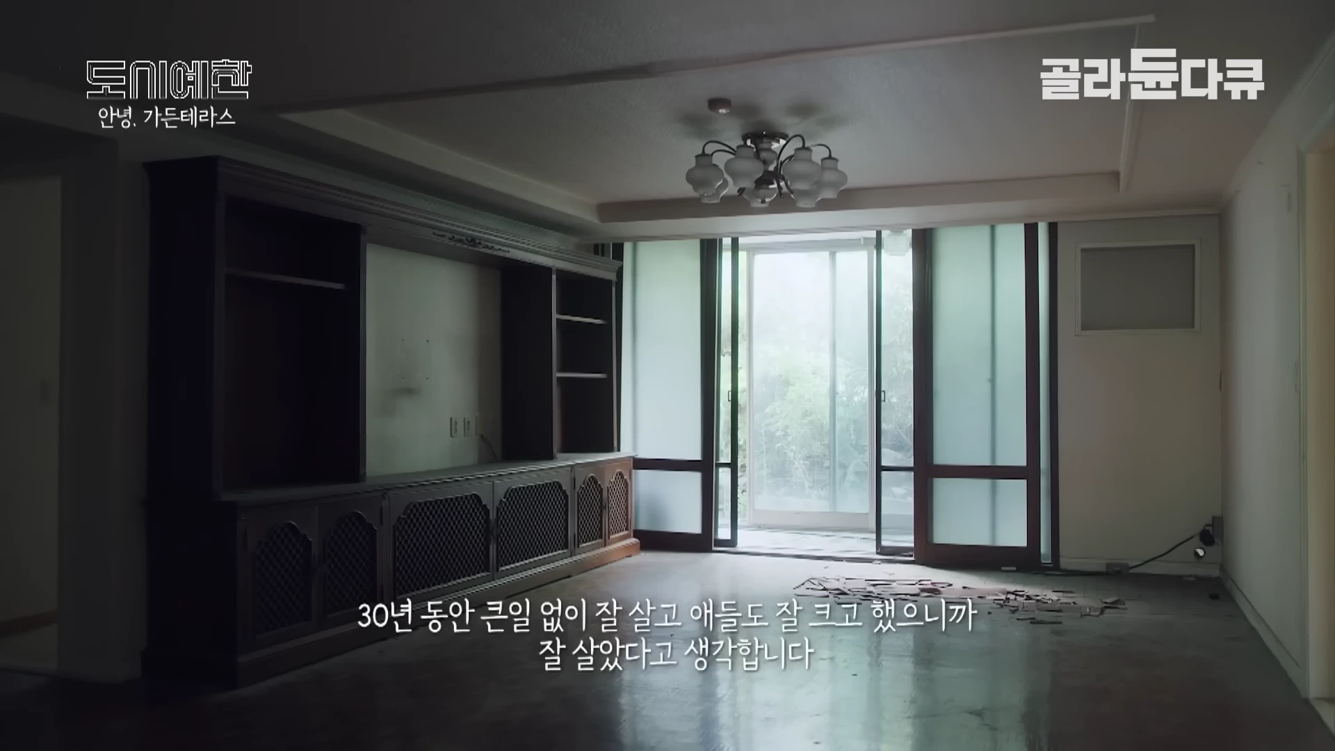 비어 있는 아파트 거실 내부의 모습으로, 한쪽 벽면에는 커다란 나무 수납장이 설치되어 있고 창밖으로는 녹음이 보인다. 화면 하단에는 자막이 쓰여 있다.