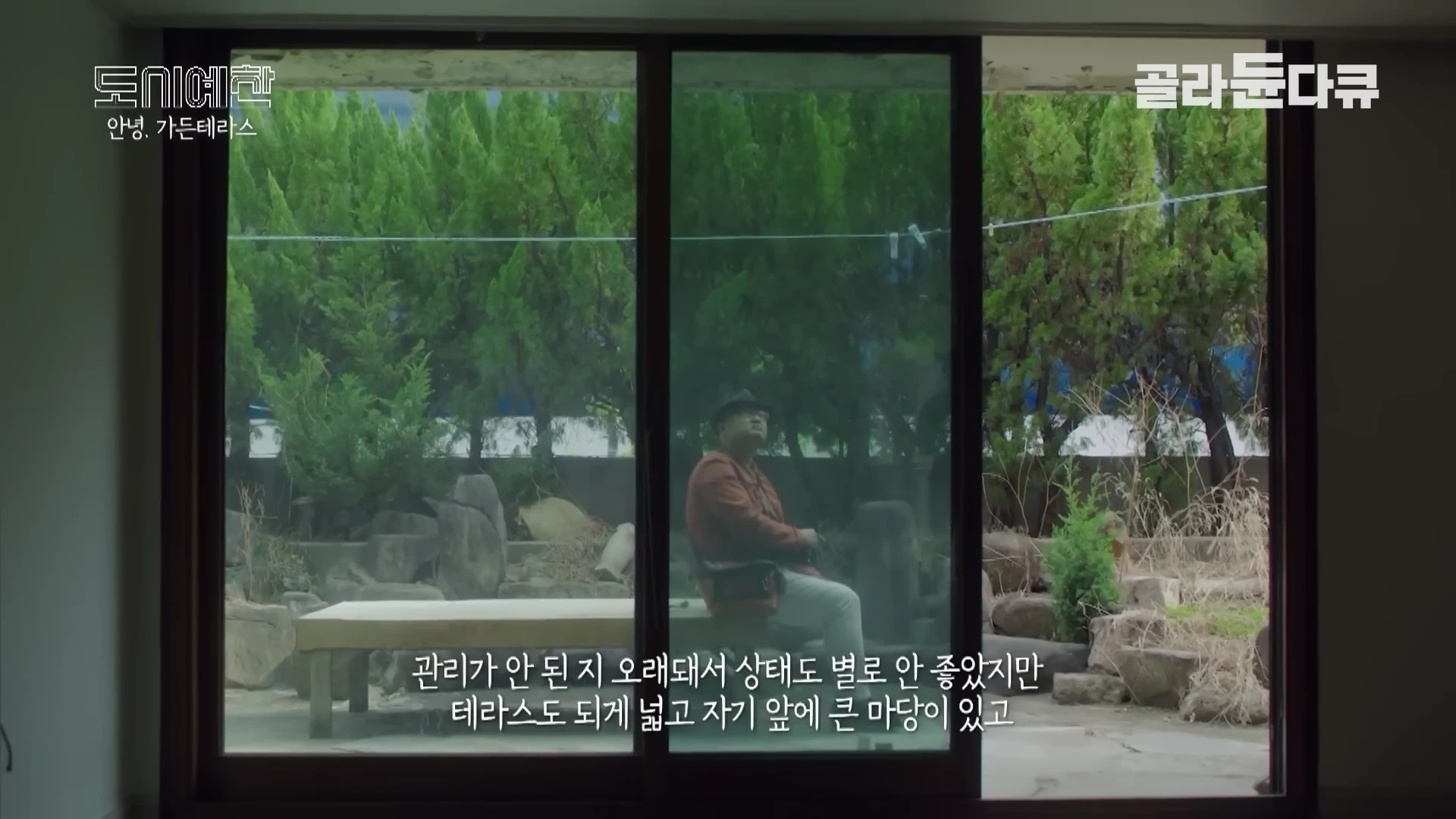 실내에서 유리창 너머로 푸른 나무와 바위가 있는 넓은 테라스 정원을 바라보는 중년 남성의 모습이 담긴 화면이다.