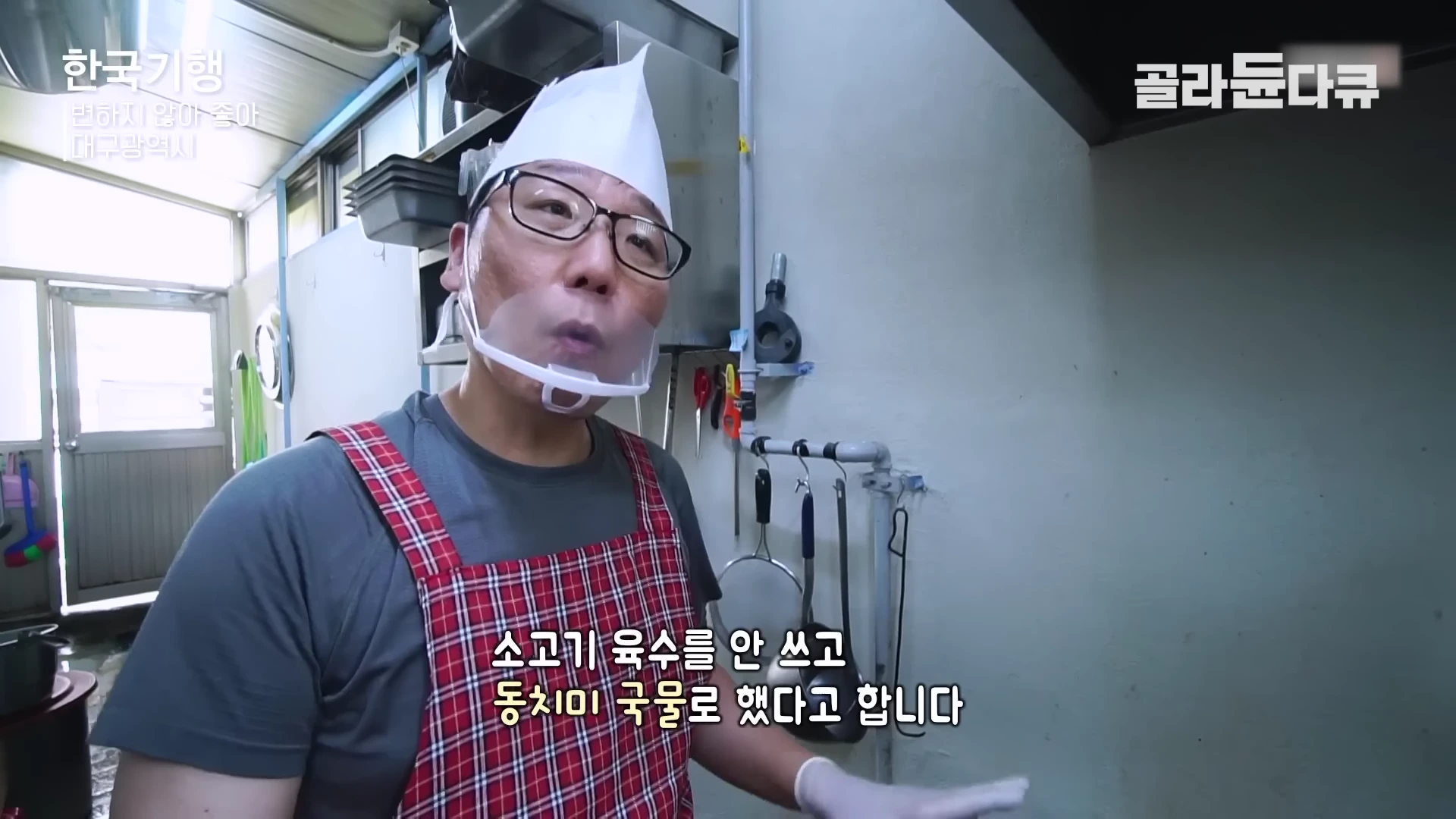 주방에서 흰색 위생 모자와 투명 마스크를 쓰고 빨간색 체크무늬 앞치마를 두른 남성이 인터뷰하는 모습이 담긴 방송 화면입니다.