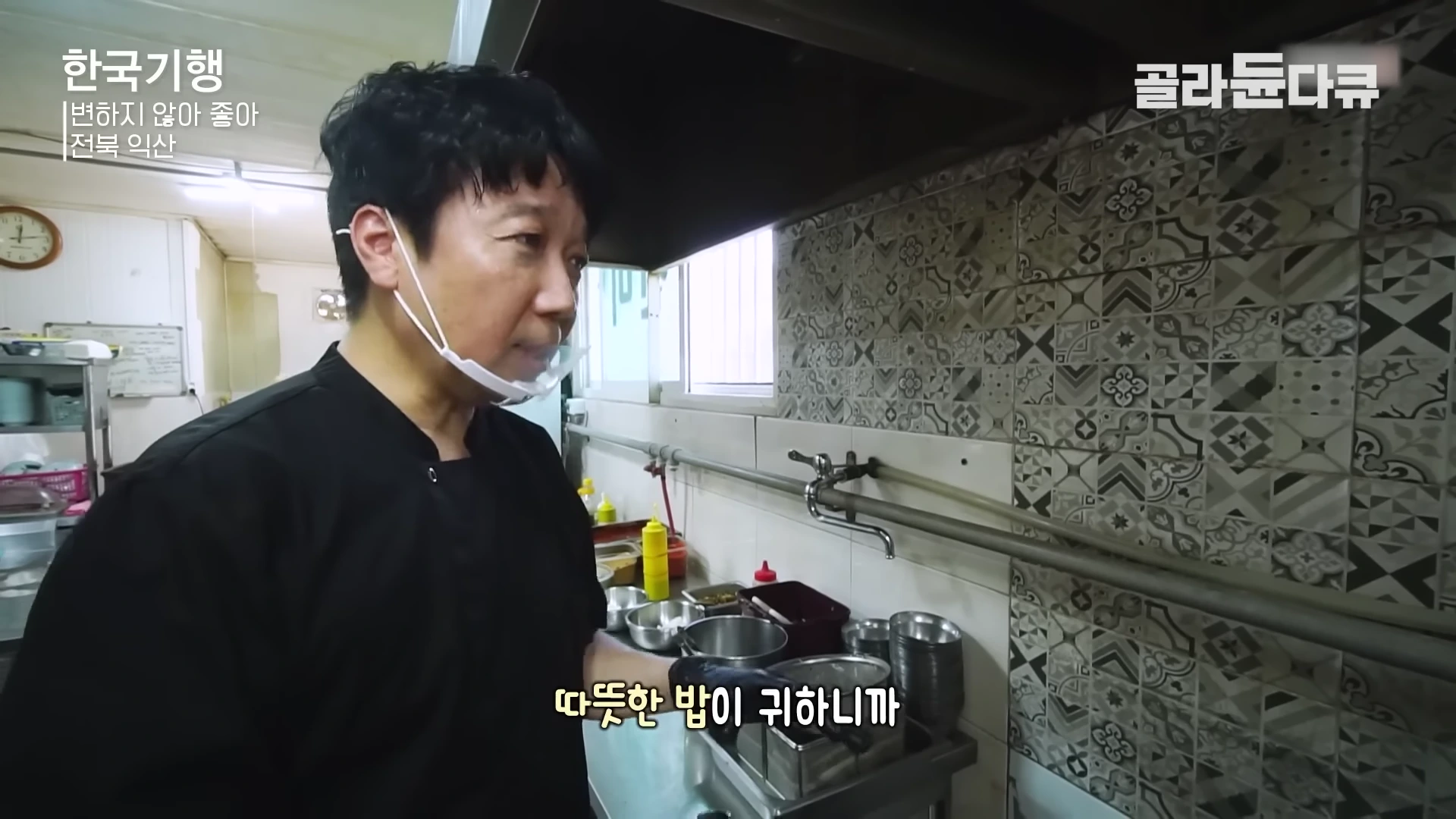 검은색 요리사 복장을 한 남성이 투명 마스크를 쓰고 주방에서 비빔밥 재료를 조리하는 모습.