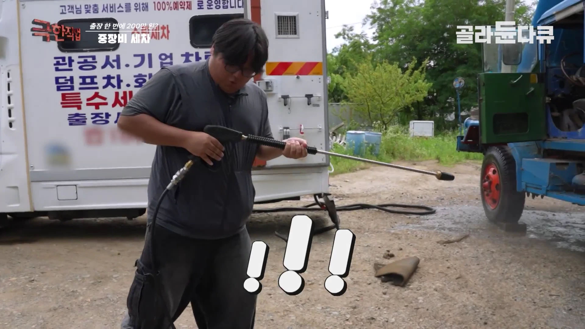 작업자가 야외에서 고압 세척기 노즐을 두 손으로 꽉 잡고 조준하고 있으며, 뒤쪽으로는 출장 세차 차량과 낡은 트럭 일부가 보인다. 화면 하단에는 놀람을 나타내는 느낌표 그래픽이 삽입되어 있다.