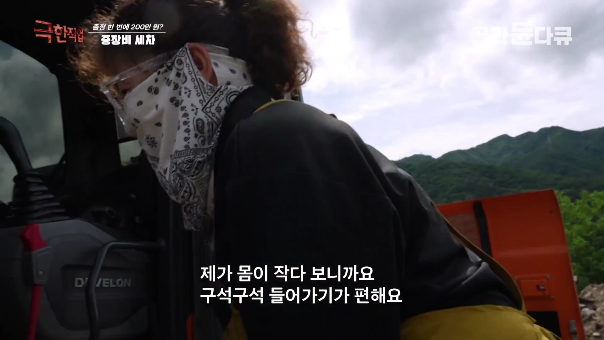 보호 안경과 두건을 착용한 작업자가 굴착기 운전석 안쪽으로 몸을 숙여 실내를 청소하고 있다.