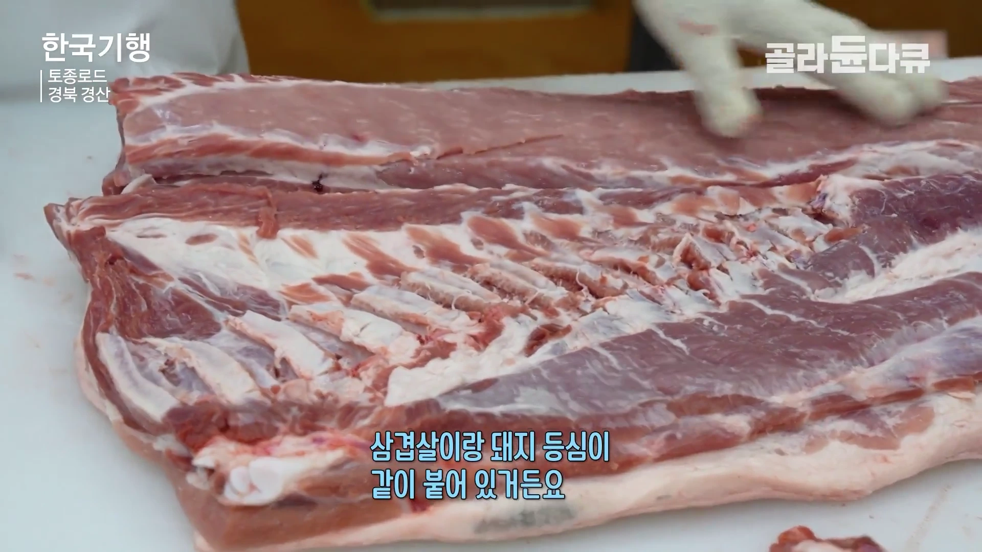 흰색 작업대 위에 놓인 큼직한 돼지고기 부위를 장갑을 낀 손이 만지며 다듬고 있는 모습