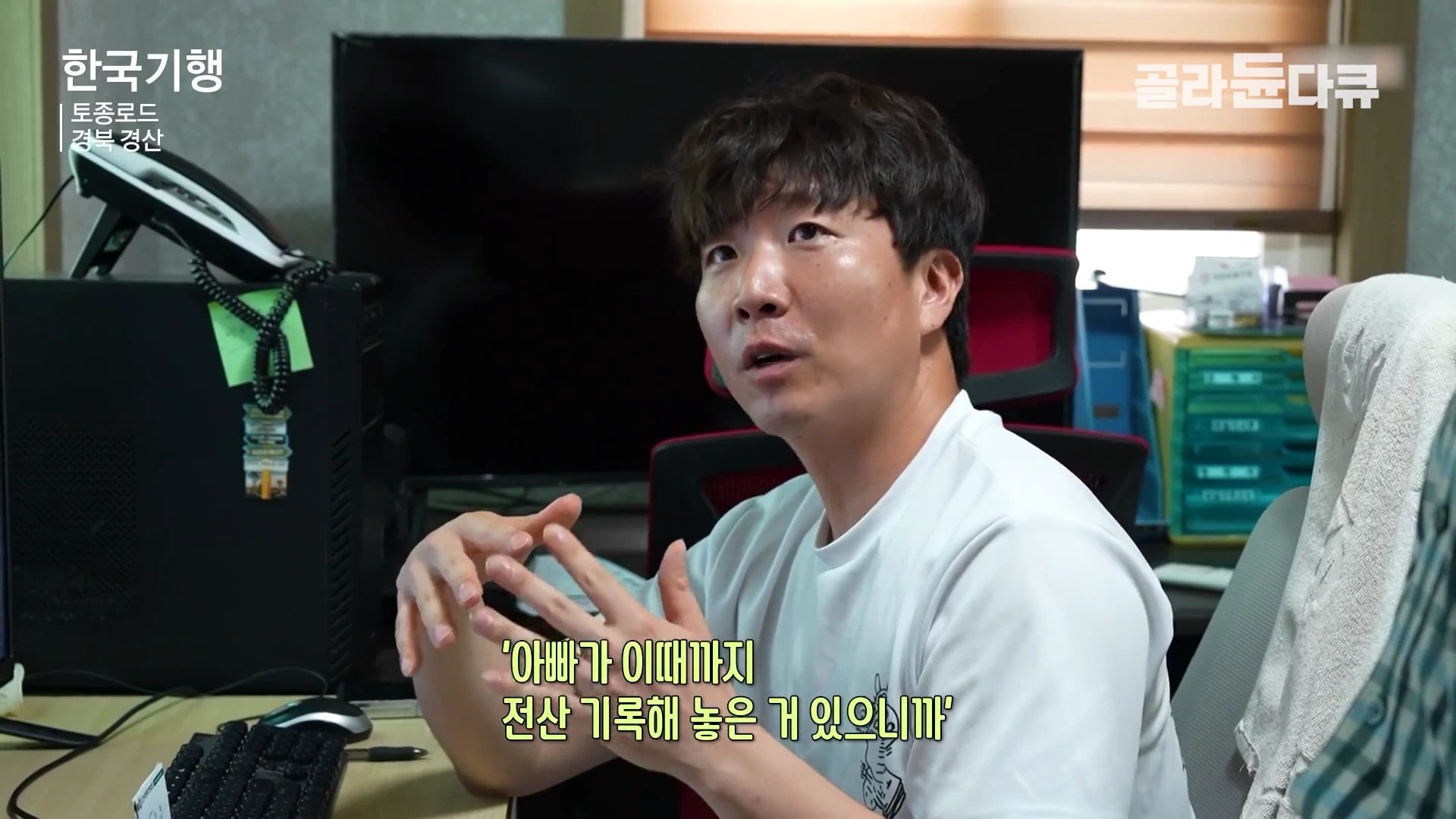 실내에서 흰색 반소매 티셔츠를 입은 젊은 남성이 책상에 앉아 대화하며 손짓하고 있고 뒤에는 컴퓨터 모니터와 전화기가 놓여 있다.