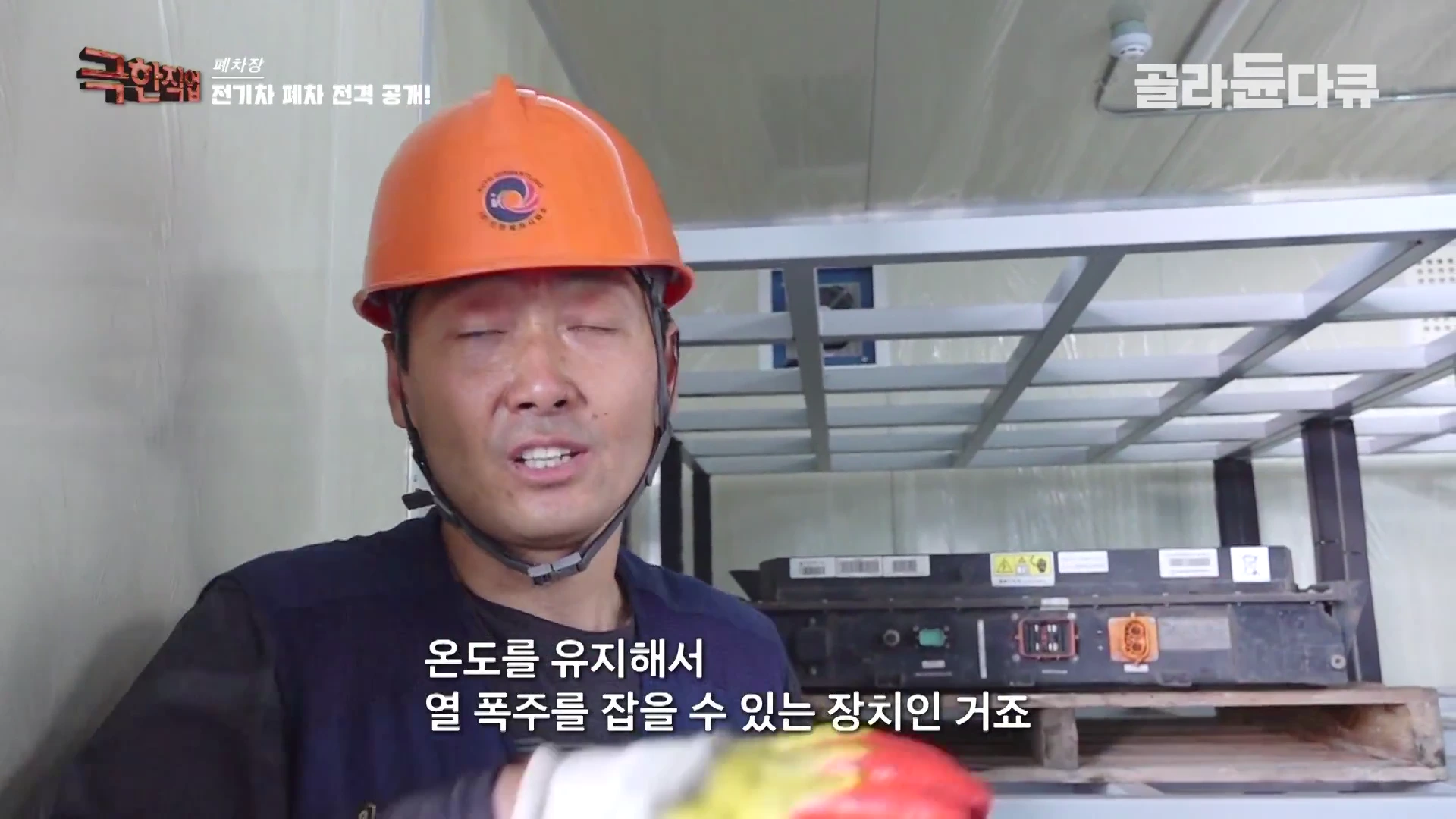 주황색 안전모를 쓴 남성이 실내 작업장에서 전기차 배터리 팩에 대해 설명하고 있는 모습