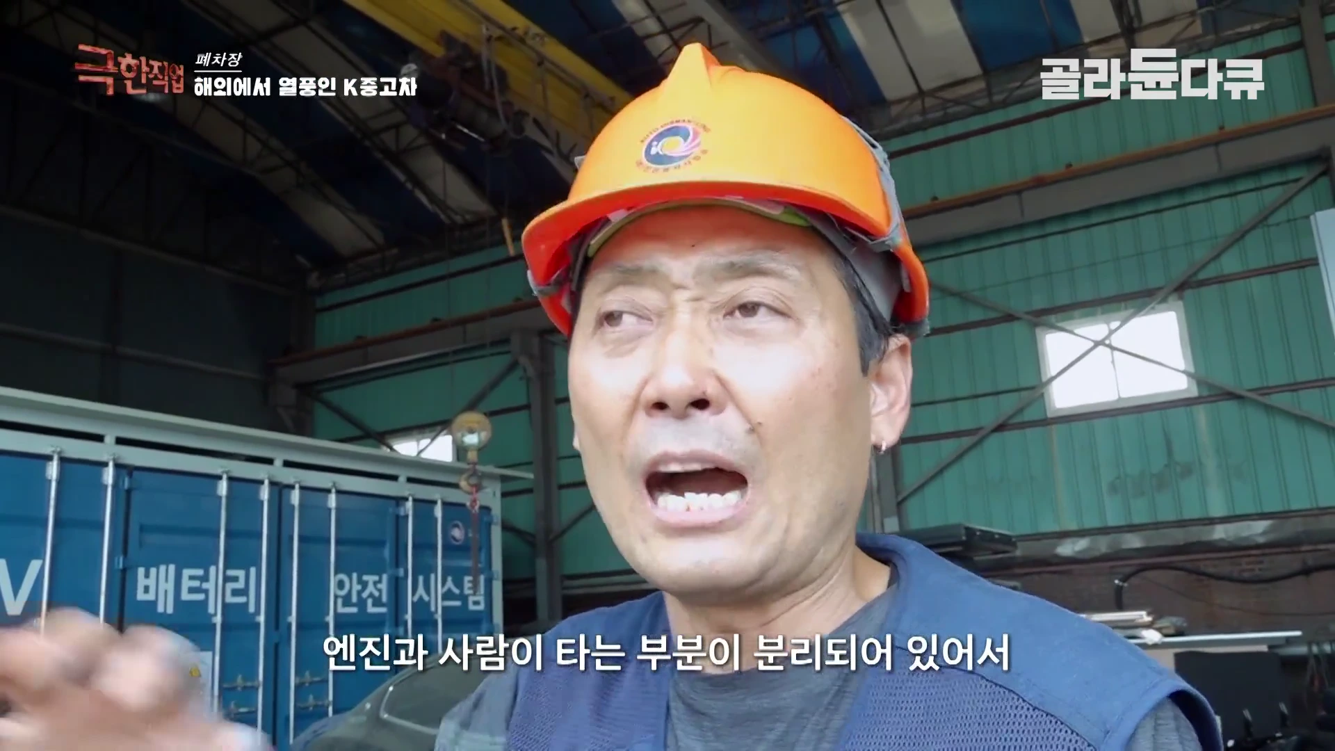 주황색 안전모를 쓴 작업자가 실내 작업장에서 이야기를 하고 있으며 배경에는 '배터리 안전 시스템'이라고 적힌 파란색 컨테이너가 보인다.