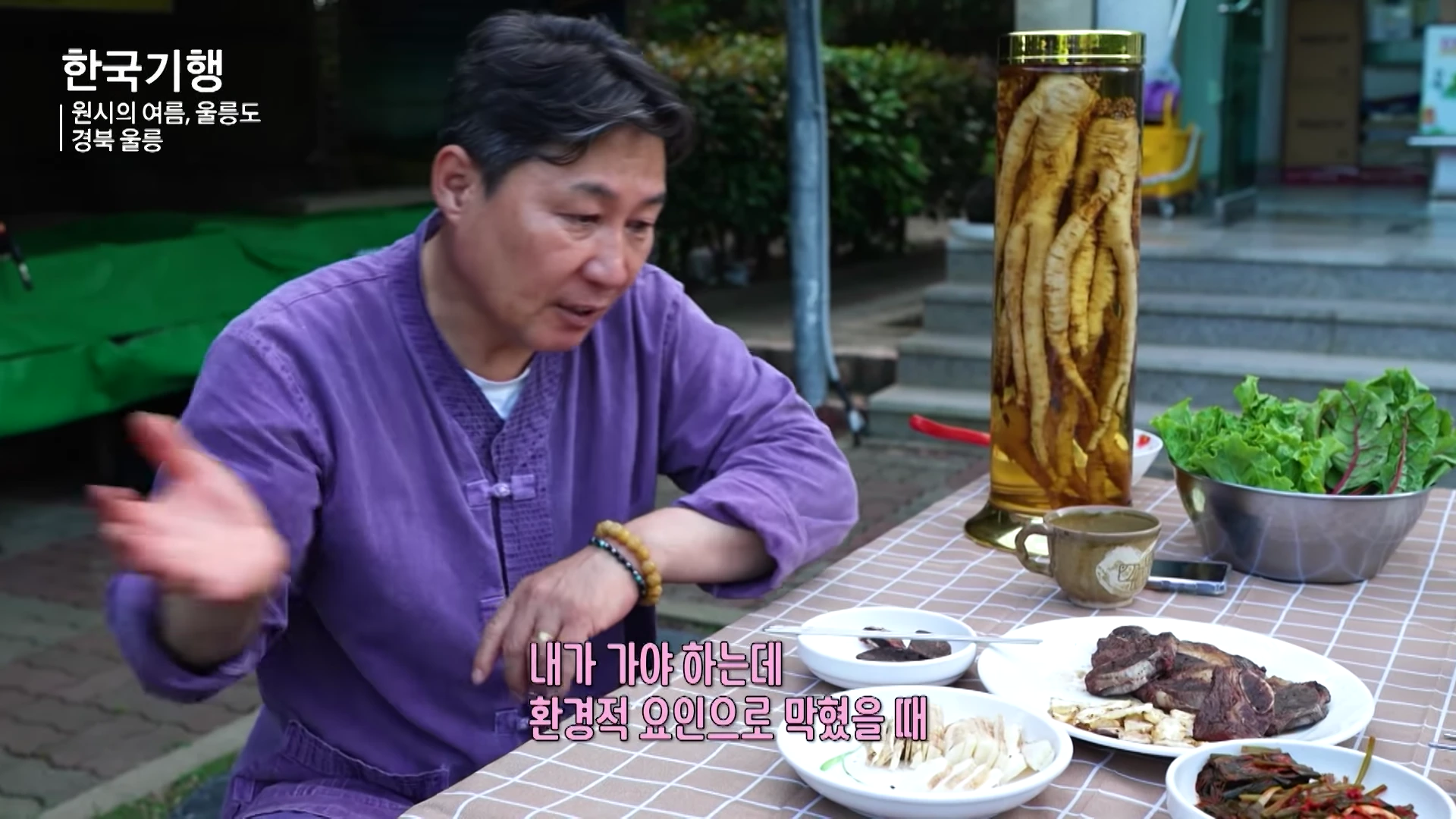 보라색 개량 한복을 입은 중년 남성이 야외 테이블에 앉아 손짓하며 대화를 나누고 있고, 테이블 위에는 구운 고기 요리와 밑반찬, 그리고 큰 인삼주 병이 놓여 있다.