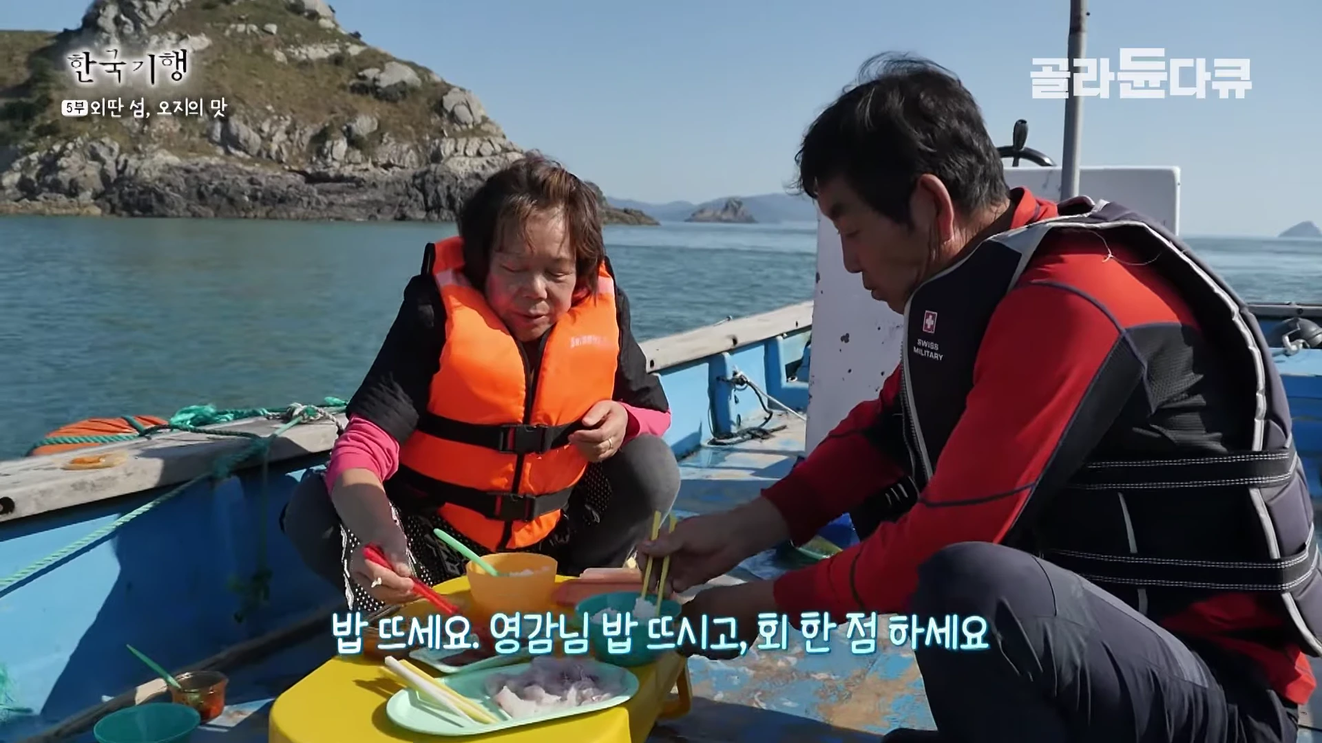 구명조끼를 입은 노부부가 작은 배 위에서 마주 앉아 회와 밥을 나누어 먹고 있는 모습이다.