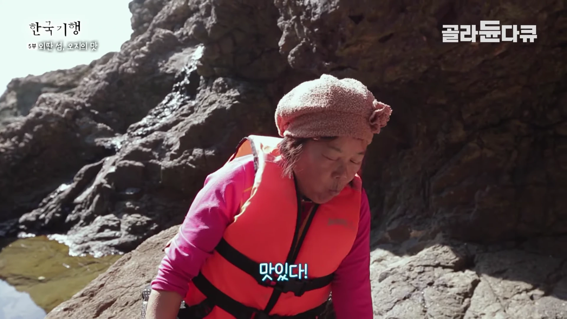 주황색 구명조끼와 베레모를 쓴 여성이 바위가 있는 해안가에서 음식을 먹으며 웃고 있는 모습