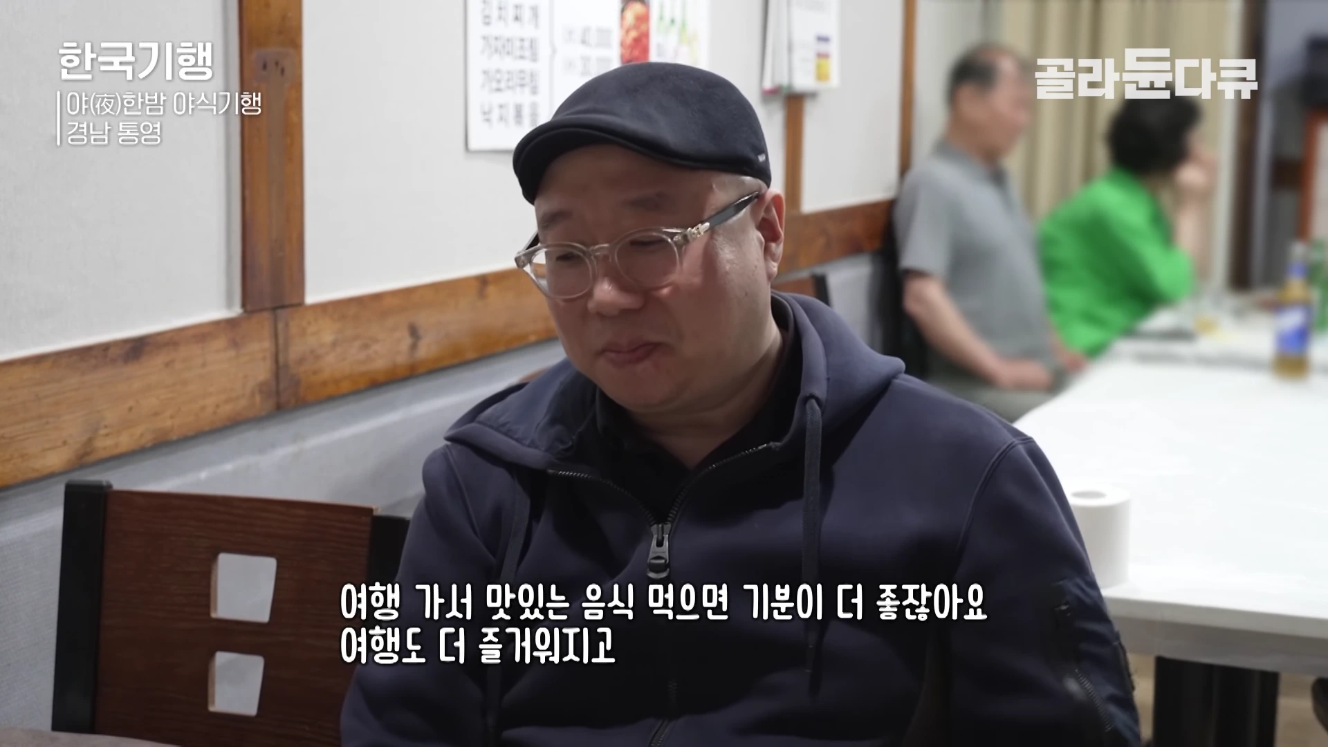 안경을 쓰고 검은색 헌팅캡을 쓴 남성이 식당 내부에서 이야기를 하고 있으며 화면 하단에는 한국어 자막이 표시되어 있습니다.