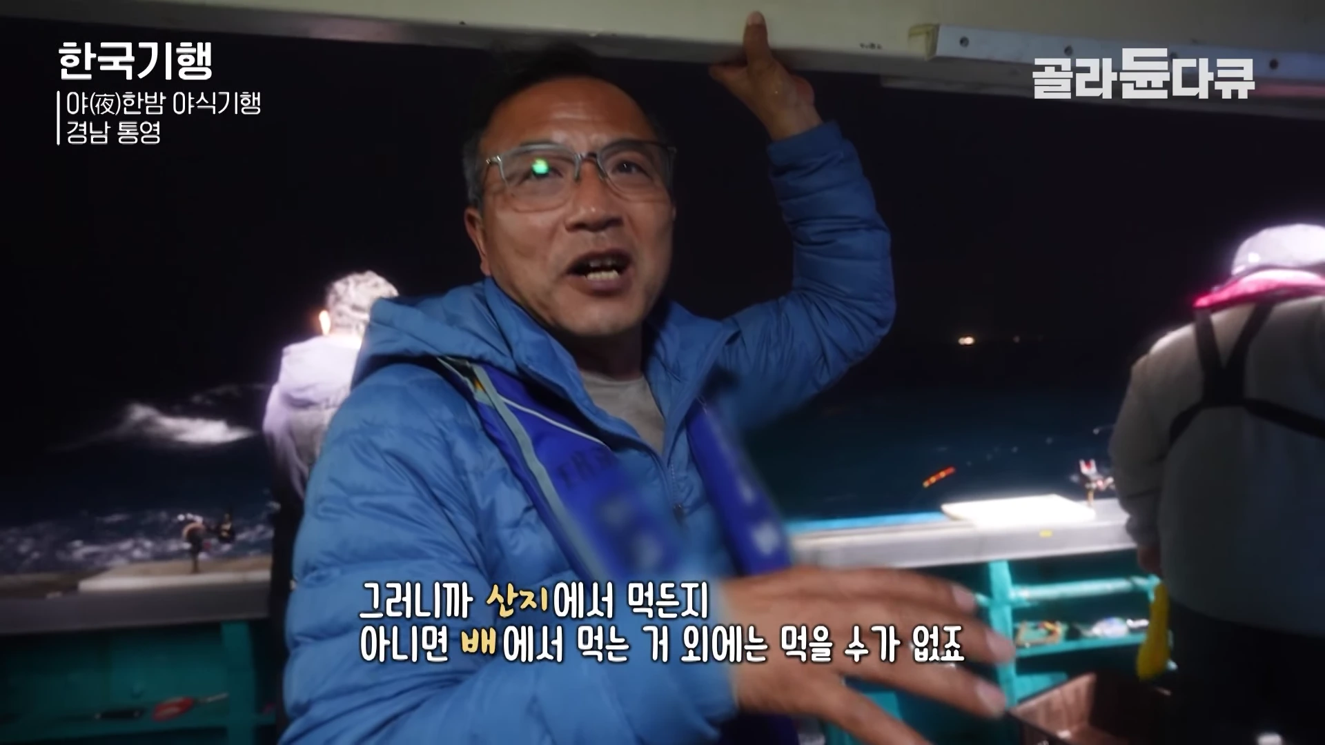 배 위에서 파란색 점퍼를 입고 안경을 쓴 남성이 환하게 웃으며 이야기하고 있으며, 배경에는 밤바다와 다른 낚시객들이 보입니다.