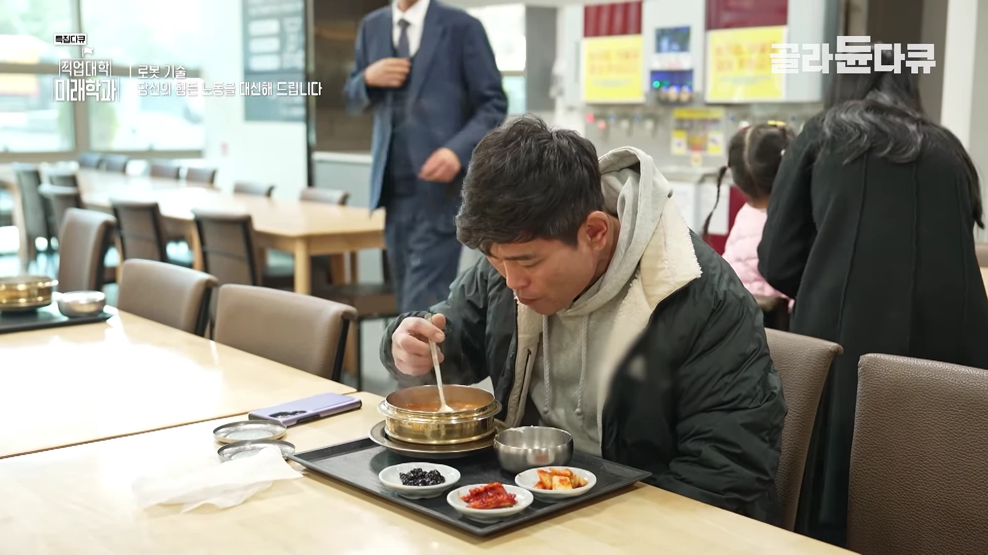 고속도로 휴게소 식당 테이블에 앉은 남성이 쟁반에 담긴 식사를 하고 있으며, 배경에는 다른 손님들과 직원이 보입니다.