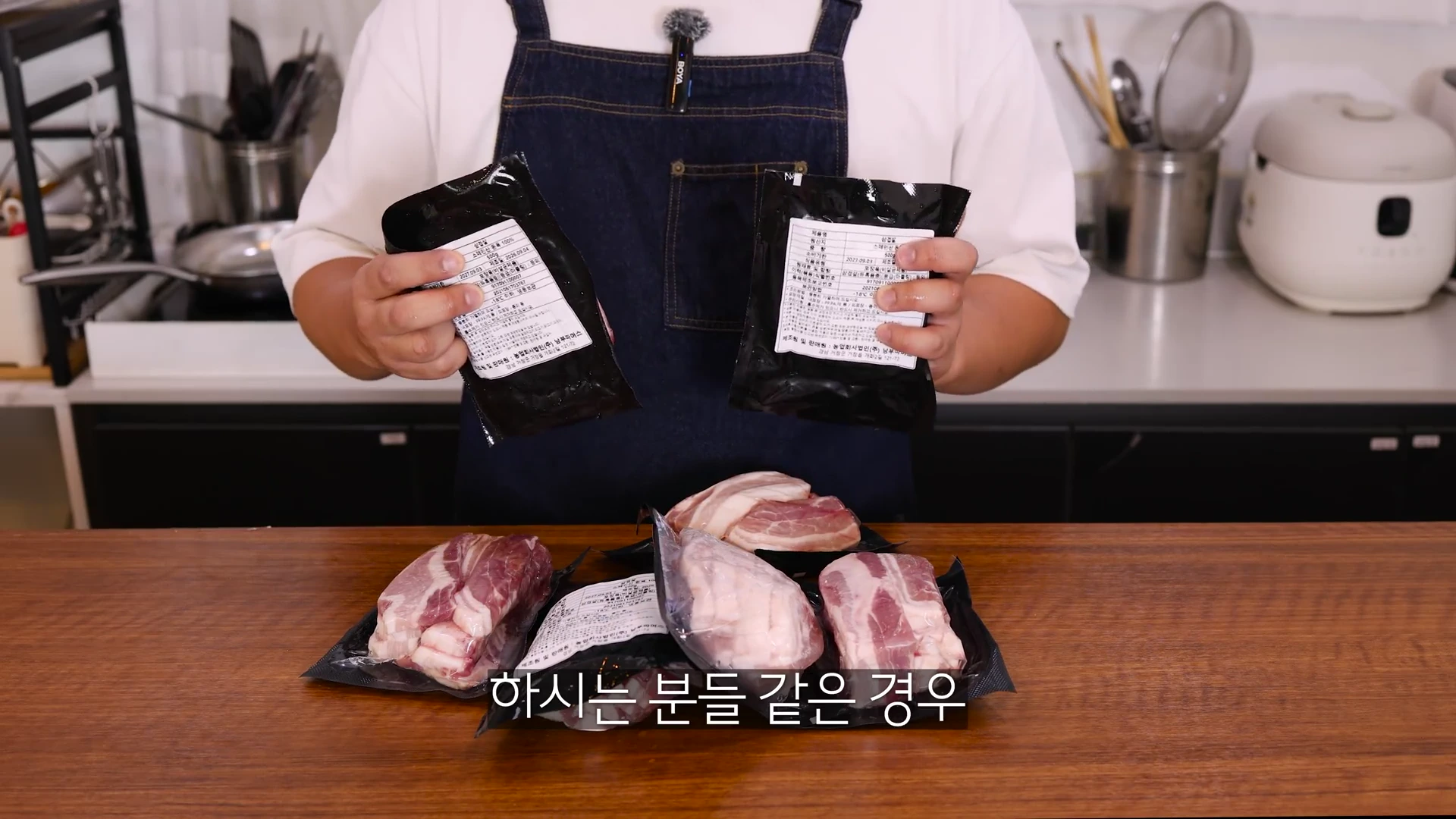 앞치마를 두른 사람이 양손에 진공 포장된 삼겹살 두 팩을 들고 있고, 테이블 위에는 포장된 고기들이 여러 개 놓여 있습니다.