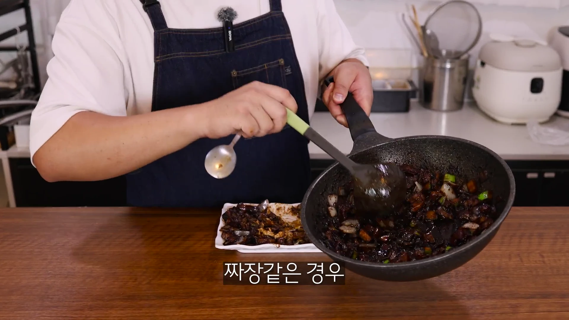 앞치마를 입은 요리사가 프라이팬에 든 춘장 소스를 국자로 덜어내고 있다.