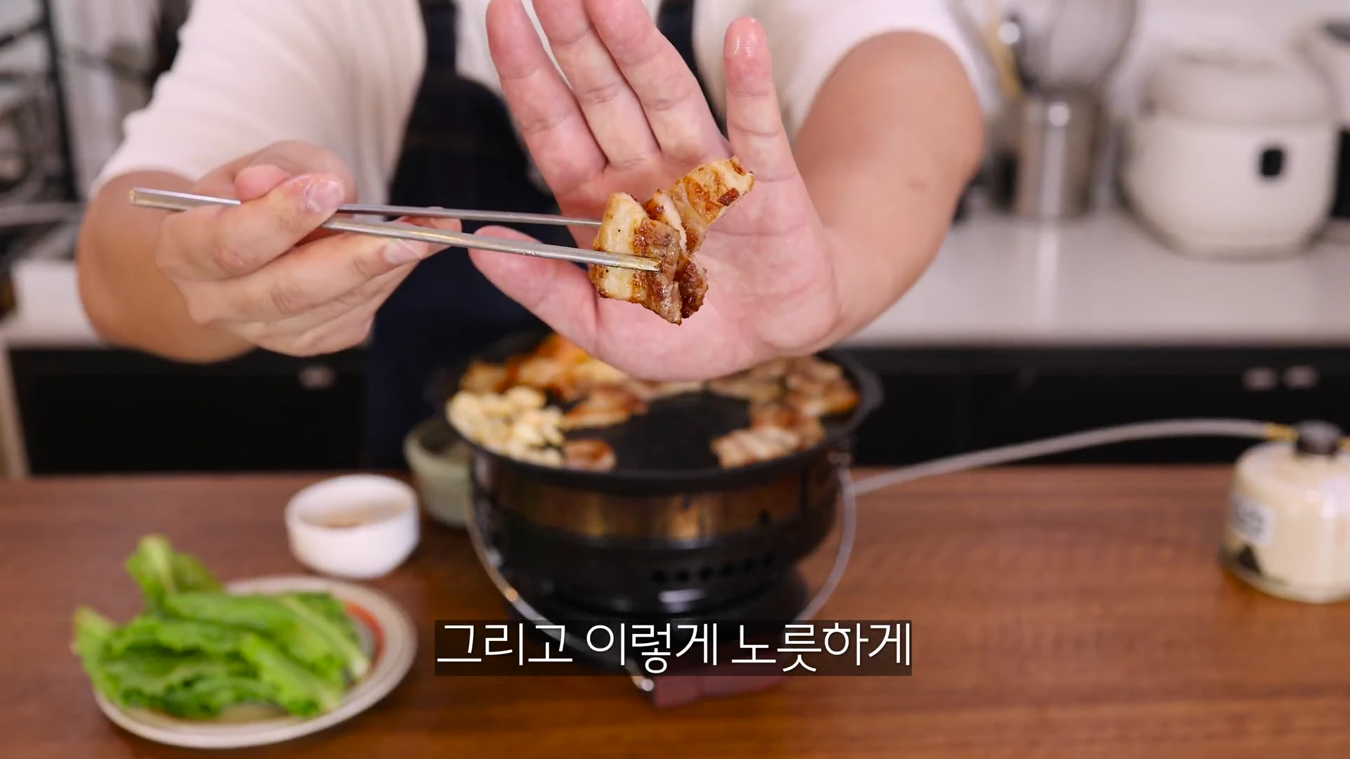 한 사람이 요리용 젓가락으로 노릇하게 구워진 삼겹살 한 점을 집어 손바닥 앞에서 보여주고 있다. 배경에는 고기가 구워지는 불판과 상추 접시가 놓여 있다.