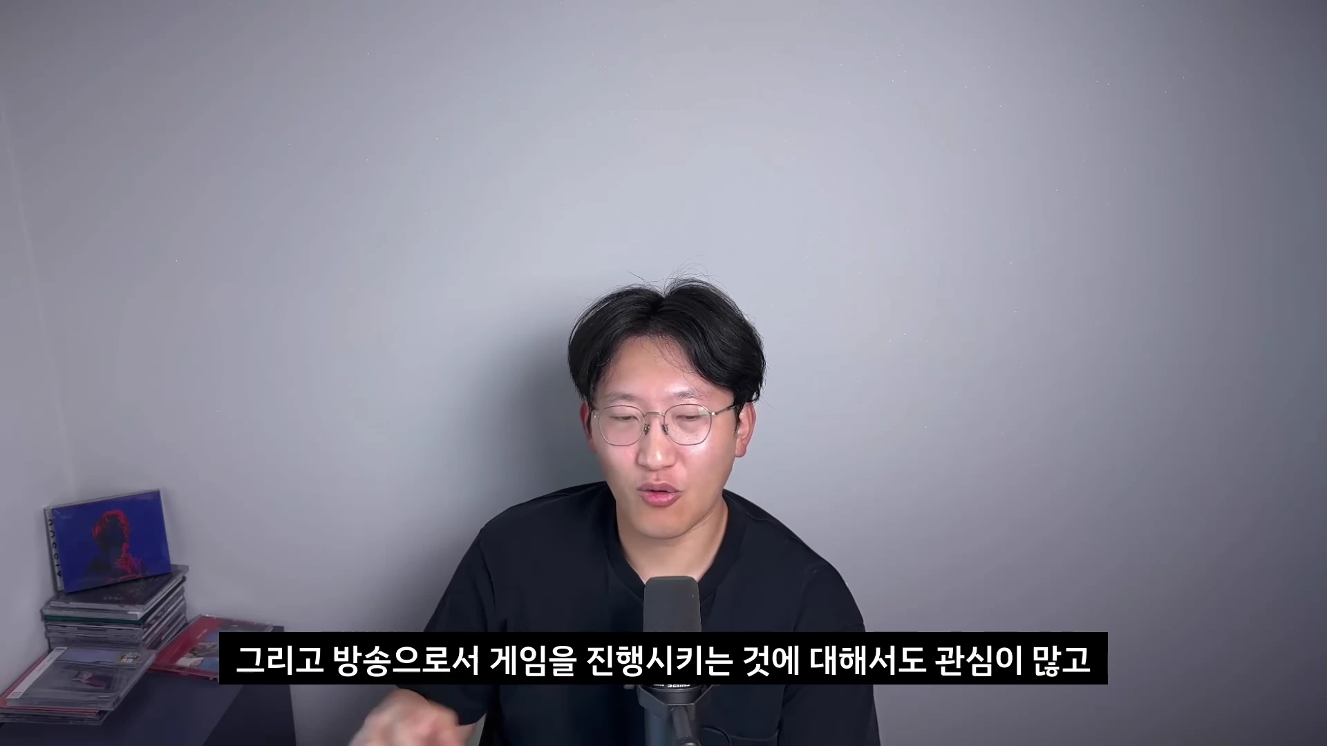 안경을 쓴 남성이 검은색 티셔츠를 입고 마이크 앞에서 이야기를 하고 있으며, 하단에는 방송 내용에 관한 한국어 자막이 표시되어 있습니다.