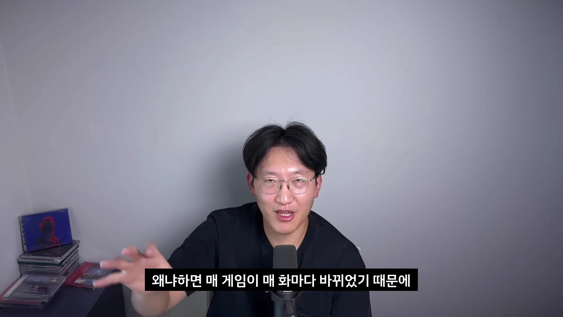 안경을 쓴 한 남성이 회색 배경 앞에서 마이크를 앞에 두고 앉아 손짓하며 이야기하고 있으며, 화면 하단에는 자막이 있습니다.