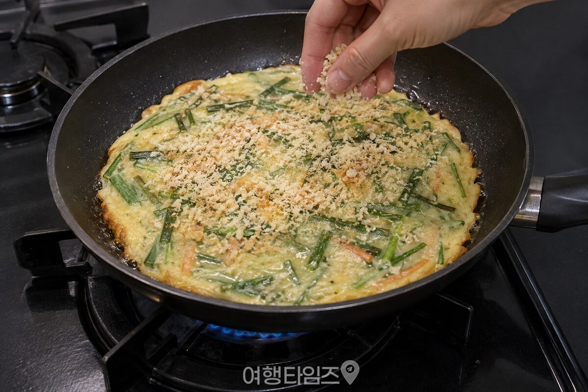 팬에서 익는 얇은 부추전의 촉촉한 윗면에 빵가루를 고르게 뿌리고 있다. / 기사 이해를 돕기 위한 생성형 이미지