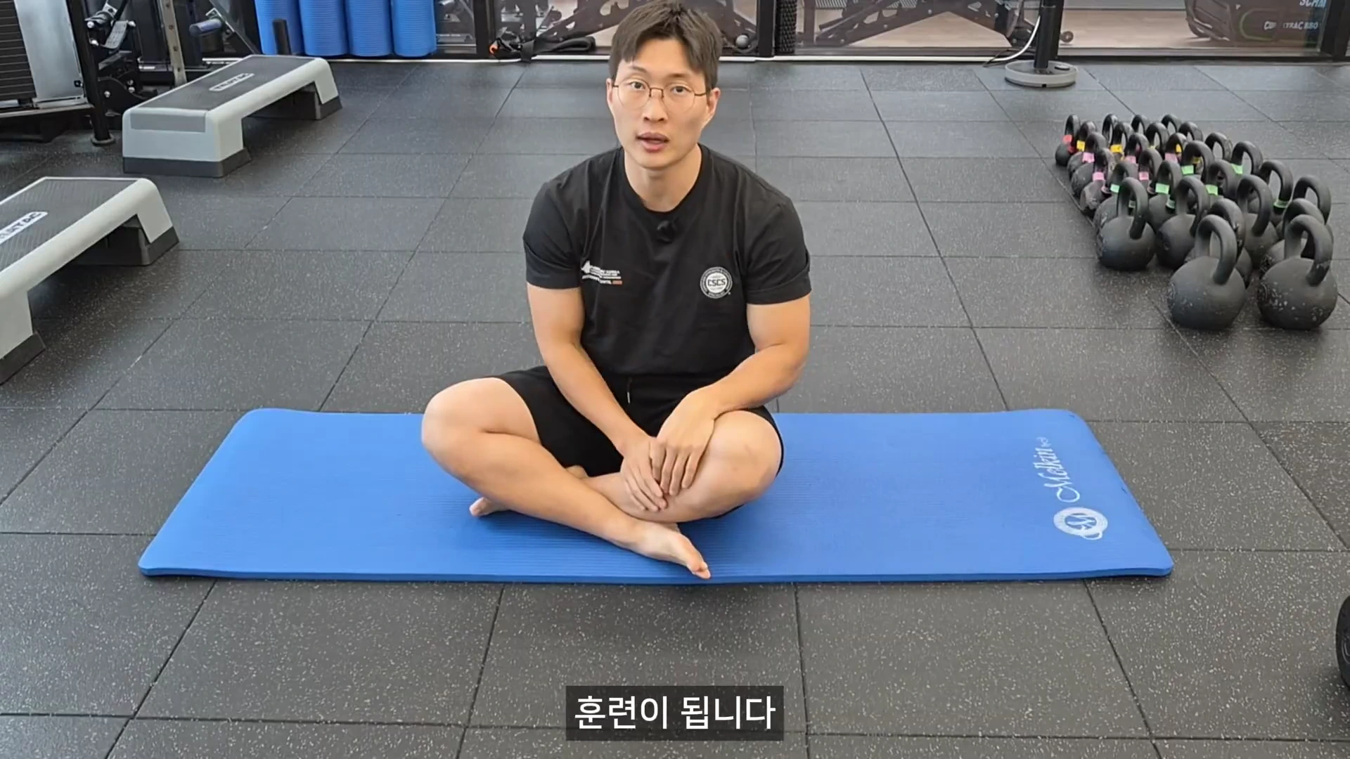 안경을 쓴 남성이 헬스장 바닥의 파란색 매트 위에서 가부좌를 틀고 앉아 카메라를 바라보며 설명하고 있다.